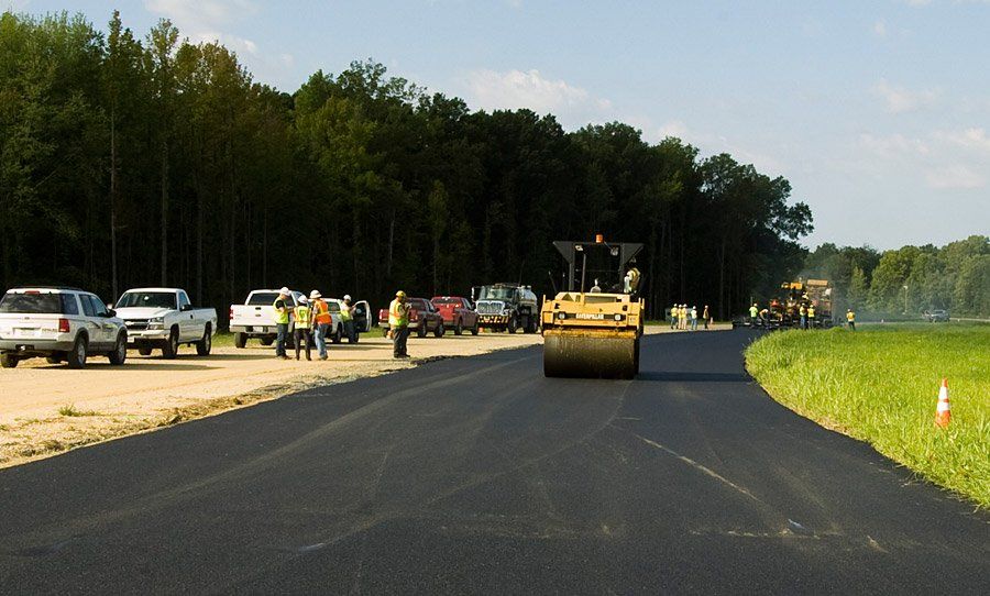 Road construction scene with a yellow road roller on fresh black asphalt and vehicles parked along the side.