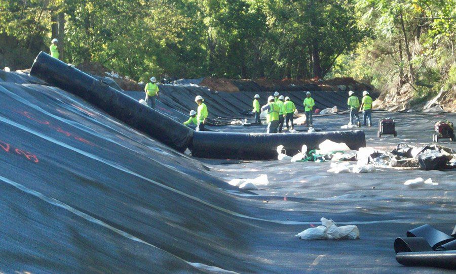 Workers in neon vests inspect a damaged bridge roadway over water, with a collapsed section in the foreground.