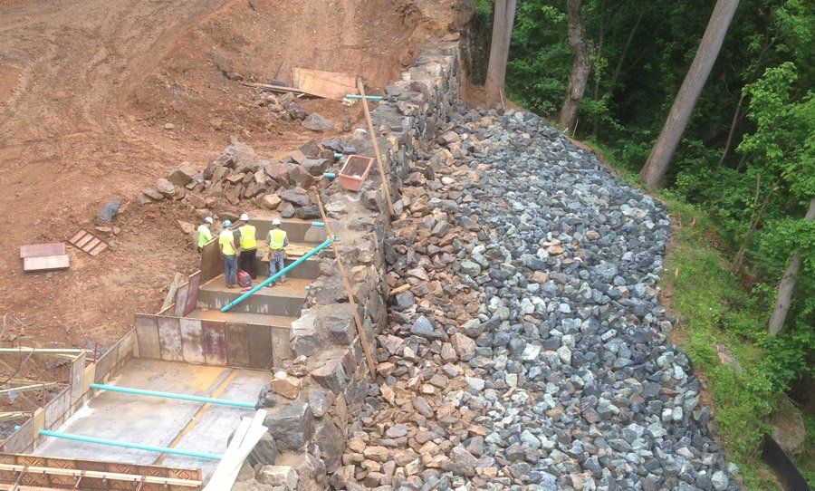 Construction workers on a rocky slope beside a retaining wall and exposed earth, with trees in the background