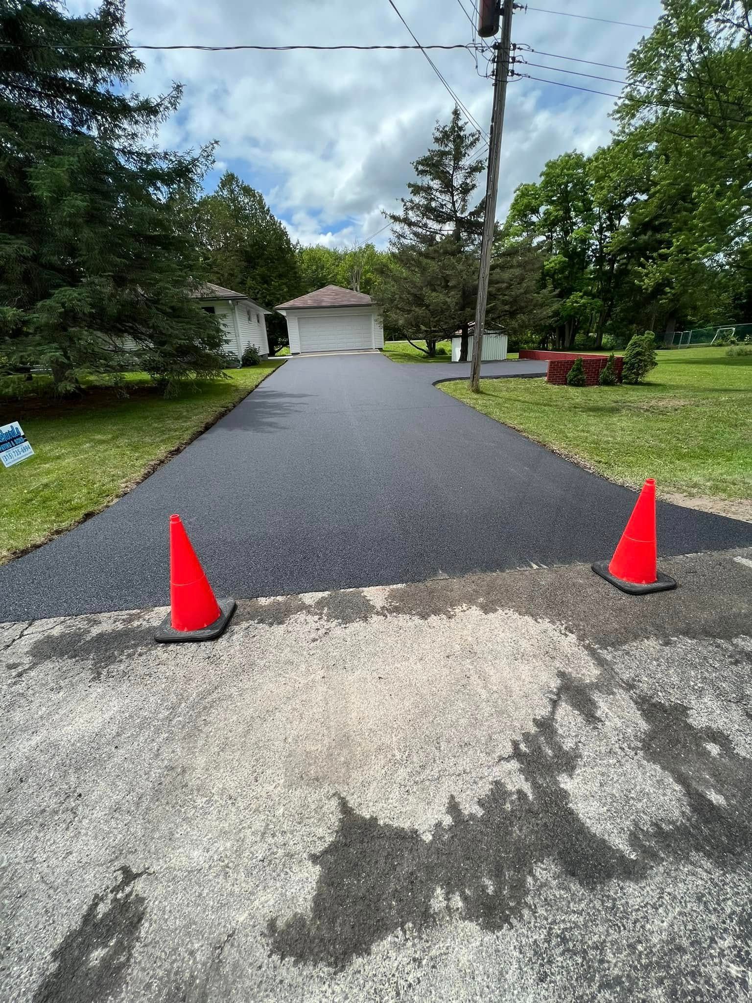 Driveway with two orange cones leading to a house under a cloudy sky, trees and grass on both sides.