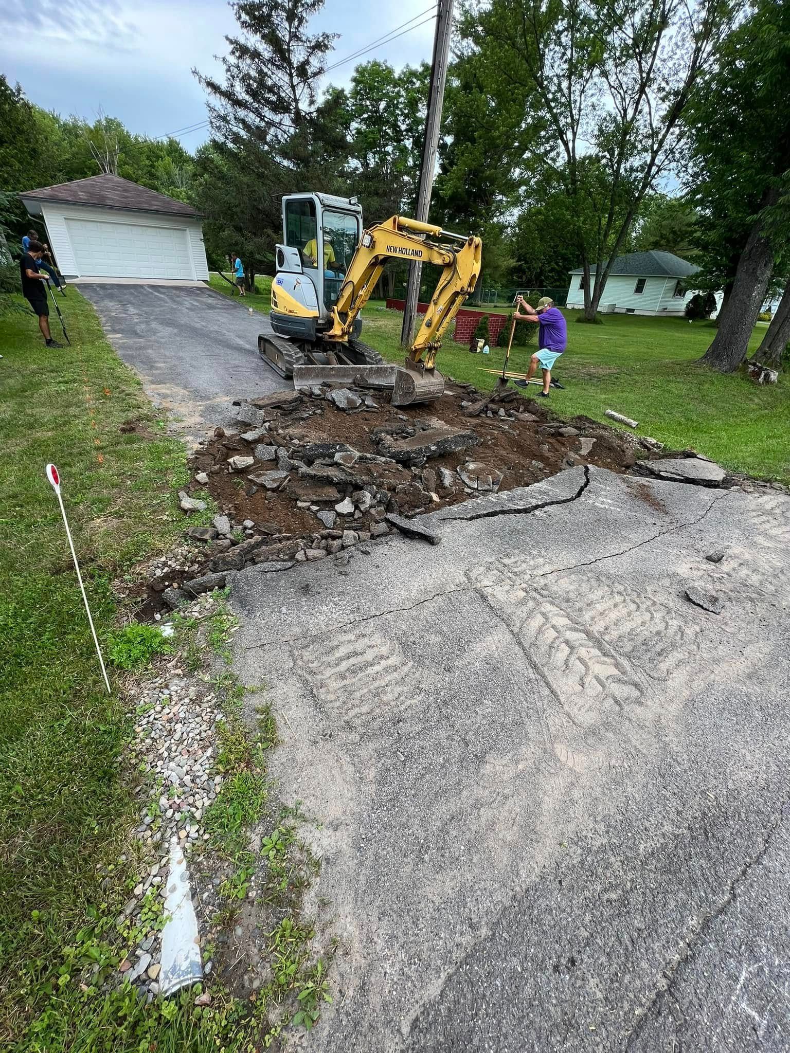 Excavator repairing a torn-up paved road near a driveway, with a worker standing nearby.