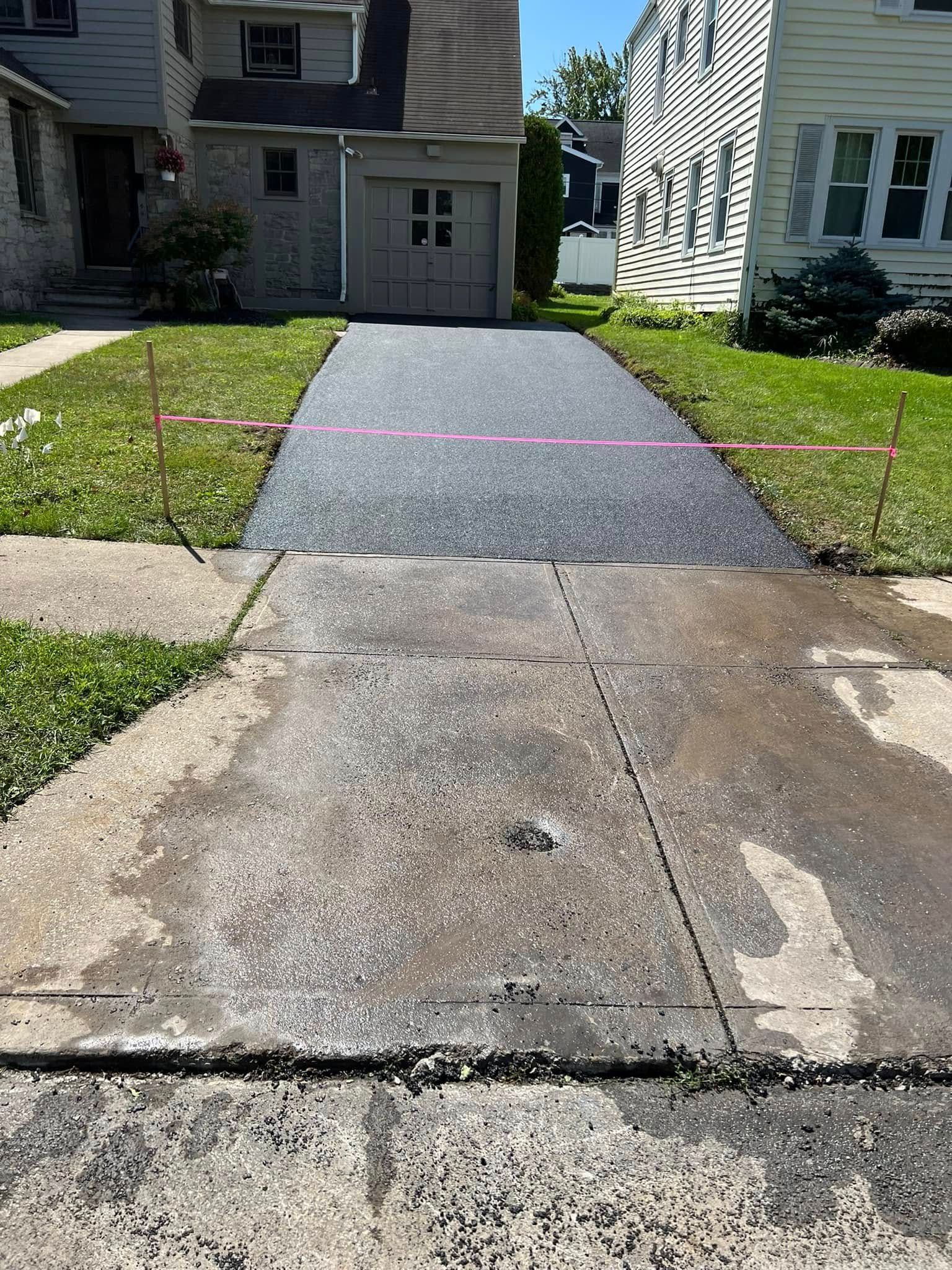 Concrete walkway leading to a house garage, with a fresh dark asphalt strip in the center.