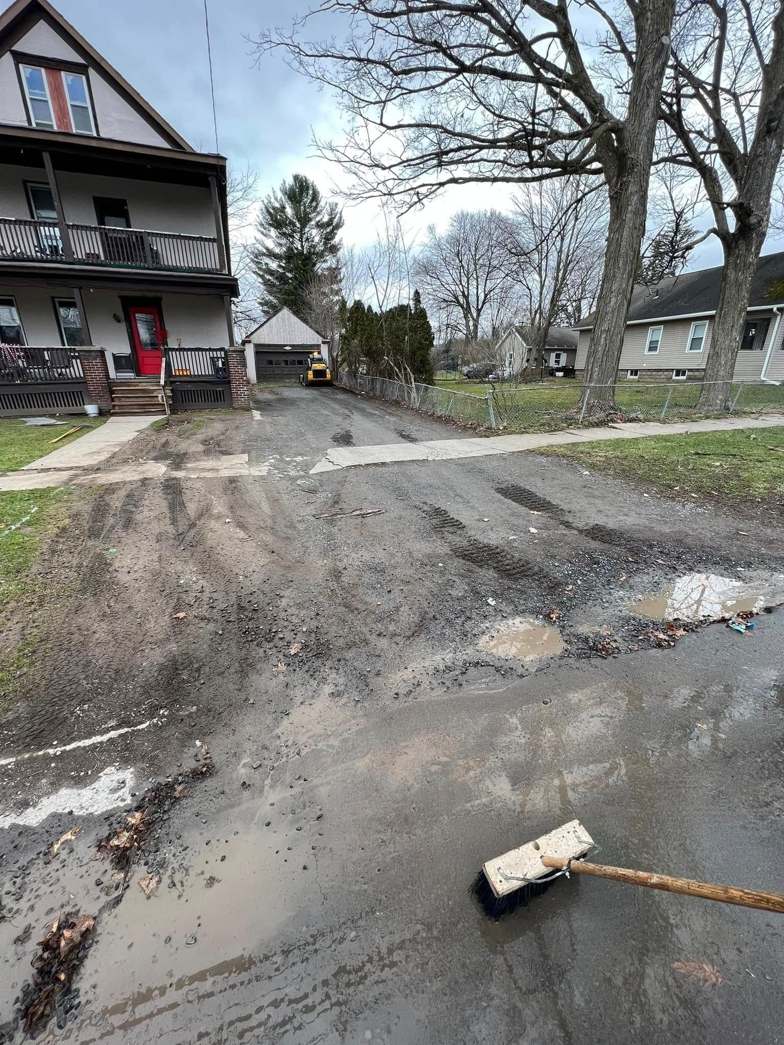 Muddy roadside with a broom in the foreground beside houses and bare trees after rain