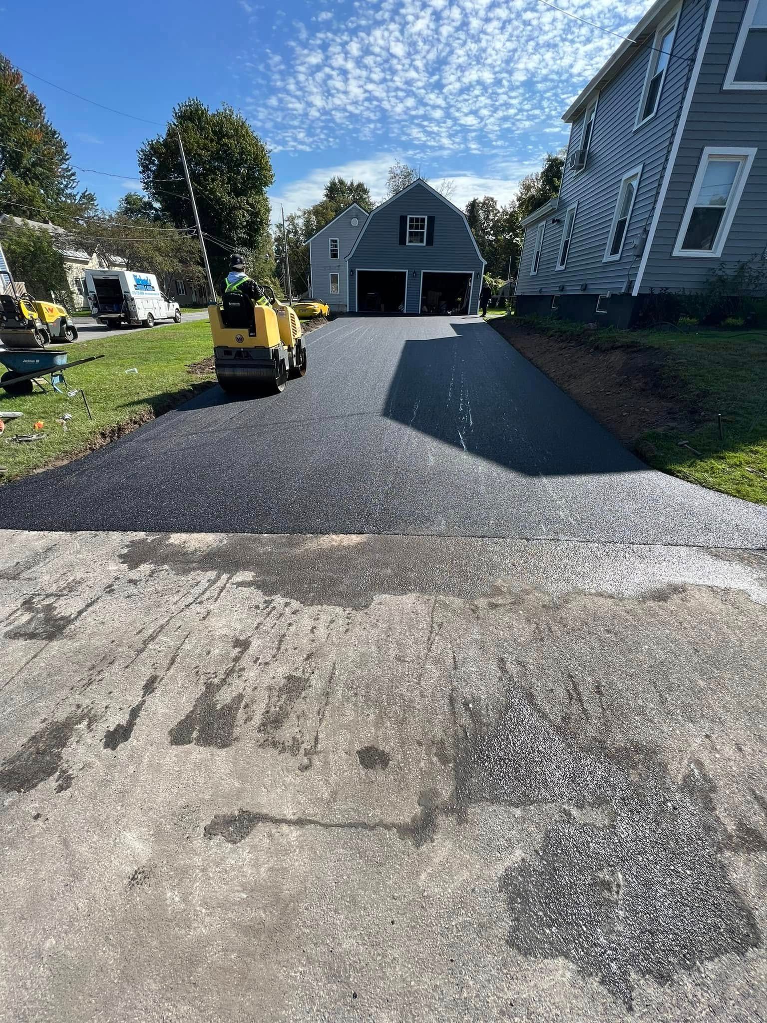 Driveway resurfacing with a yellow machine, leading to a house and garage on a sunny day