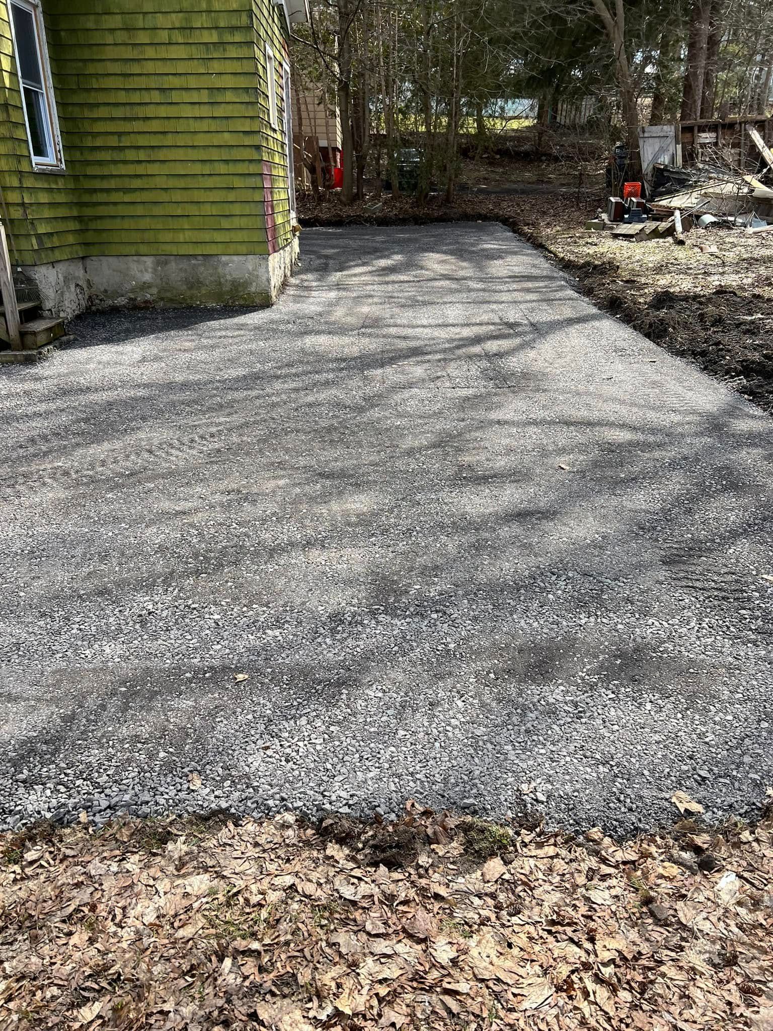 Gravel driveway beside a green building, with fallen leaves in the foreground and trees ahead.