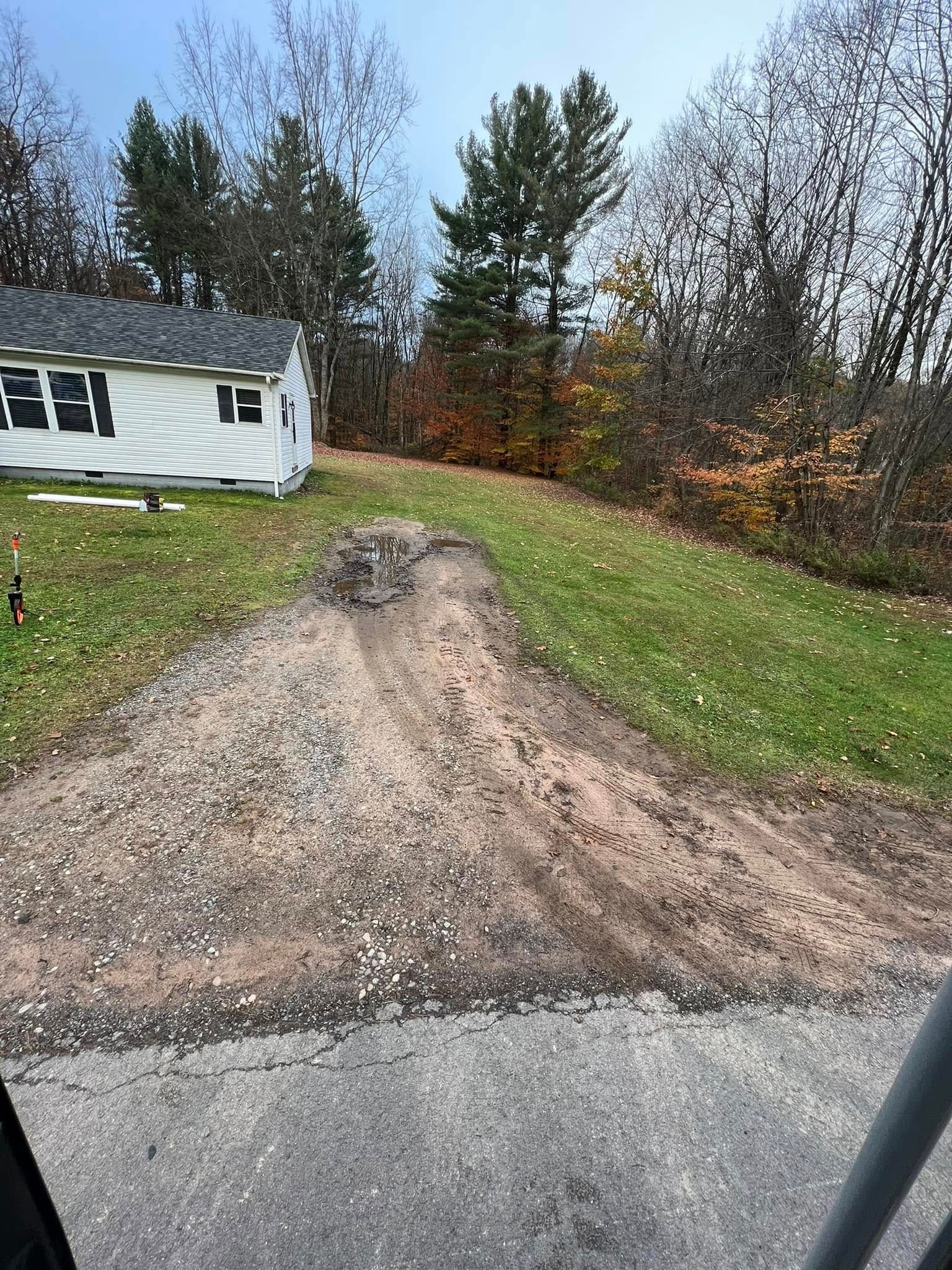 Dirt driveway leading up a grassy hill beside a white house, with autumn trees in the background.