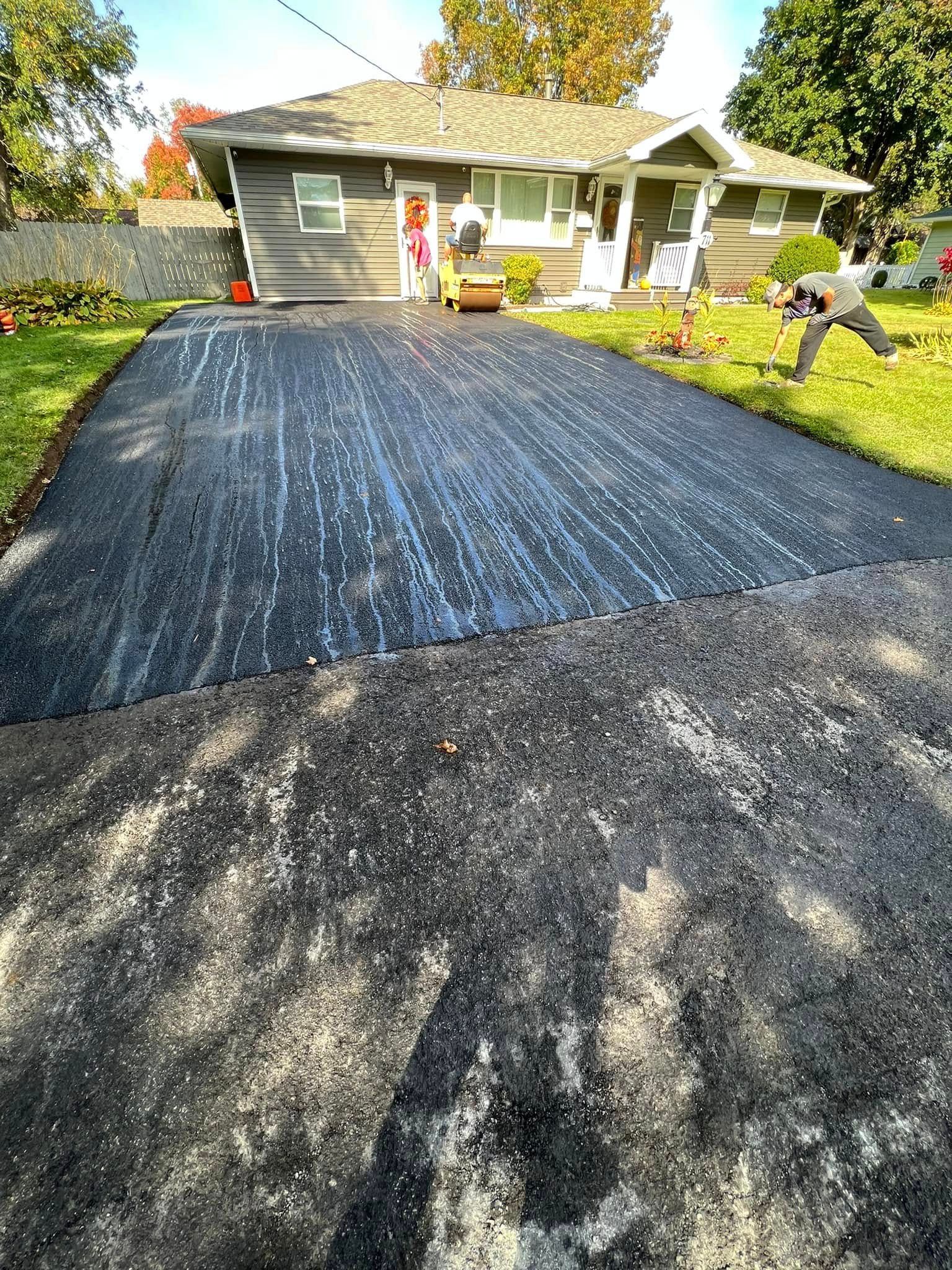 Freshly resurfaced black driveway in front of a house, with two workers on the lawn