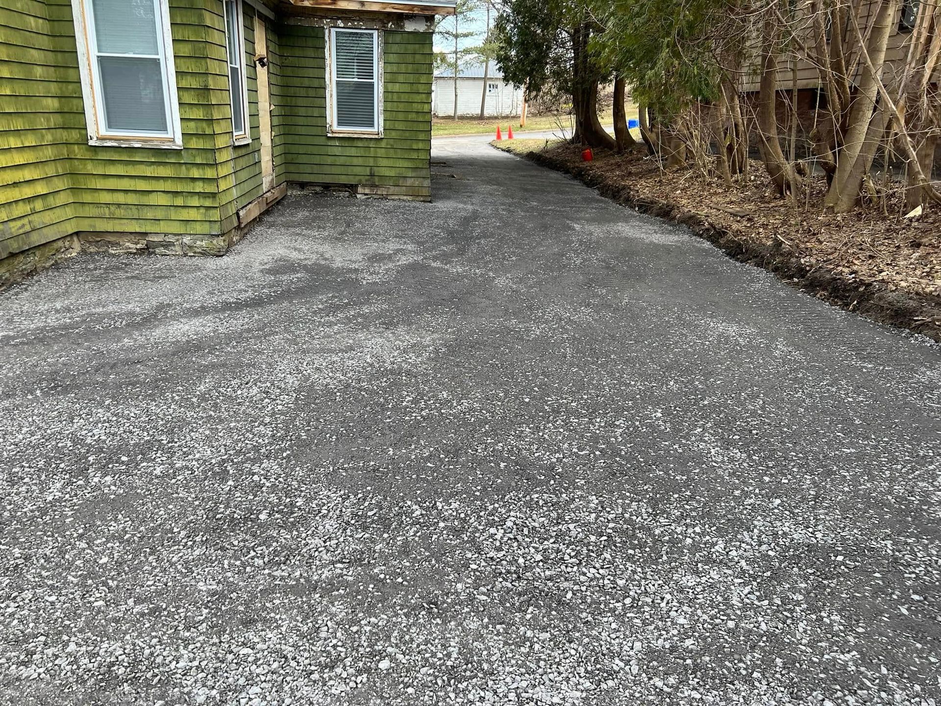 Gravel driveway beside a green house and leafless trees in late winter.