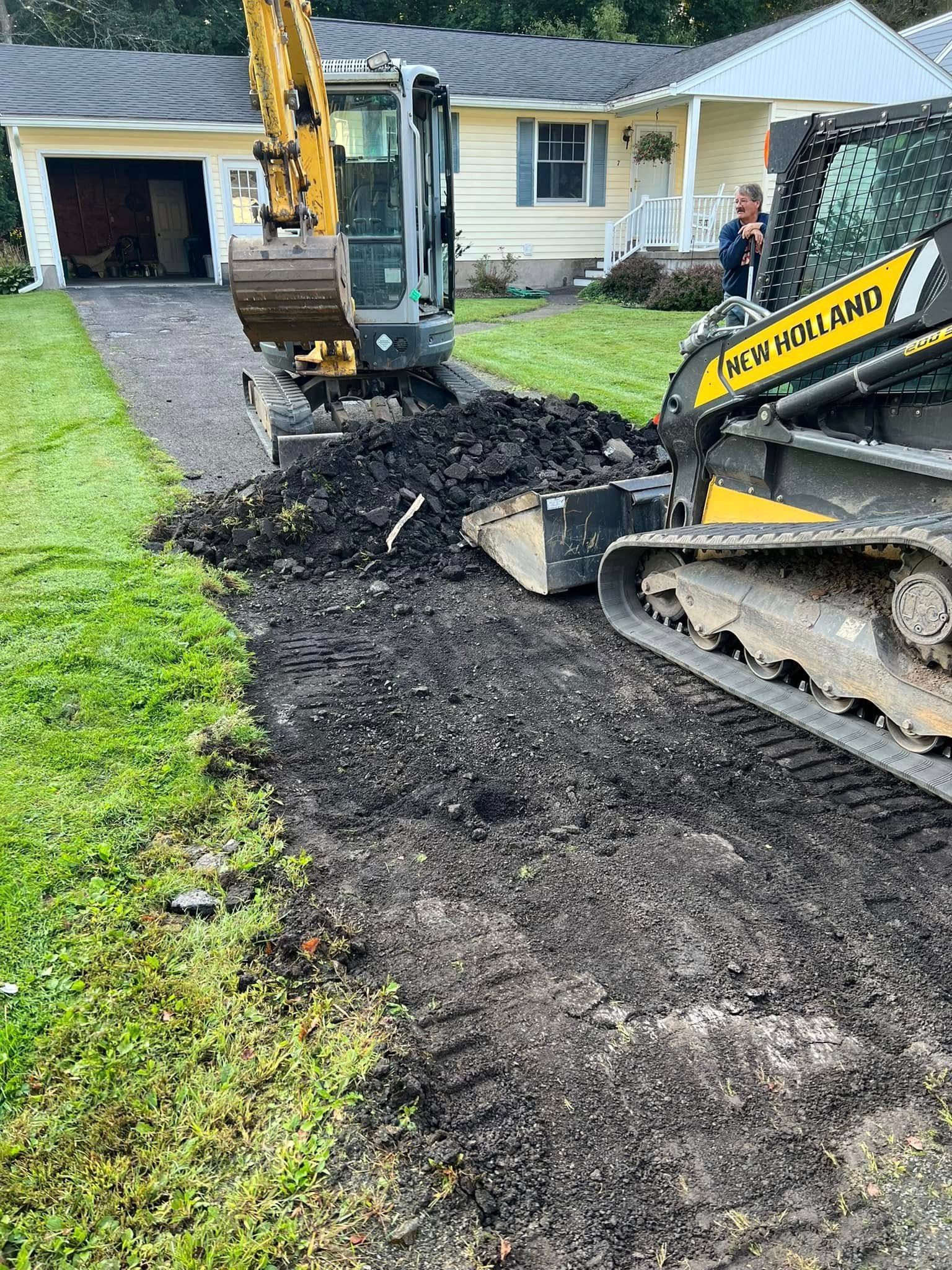 Excavator tearing up a front yard beside a yellow house, with soil piled along the driveway.