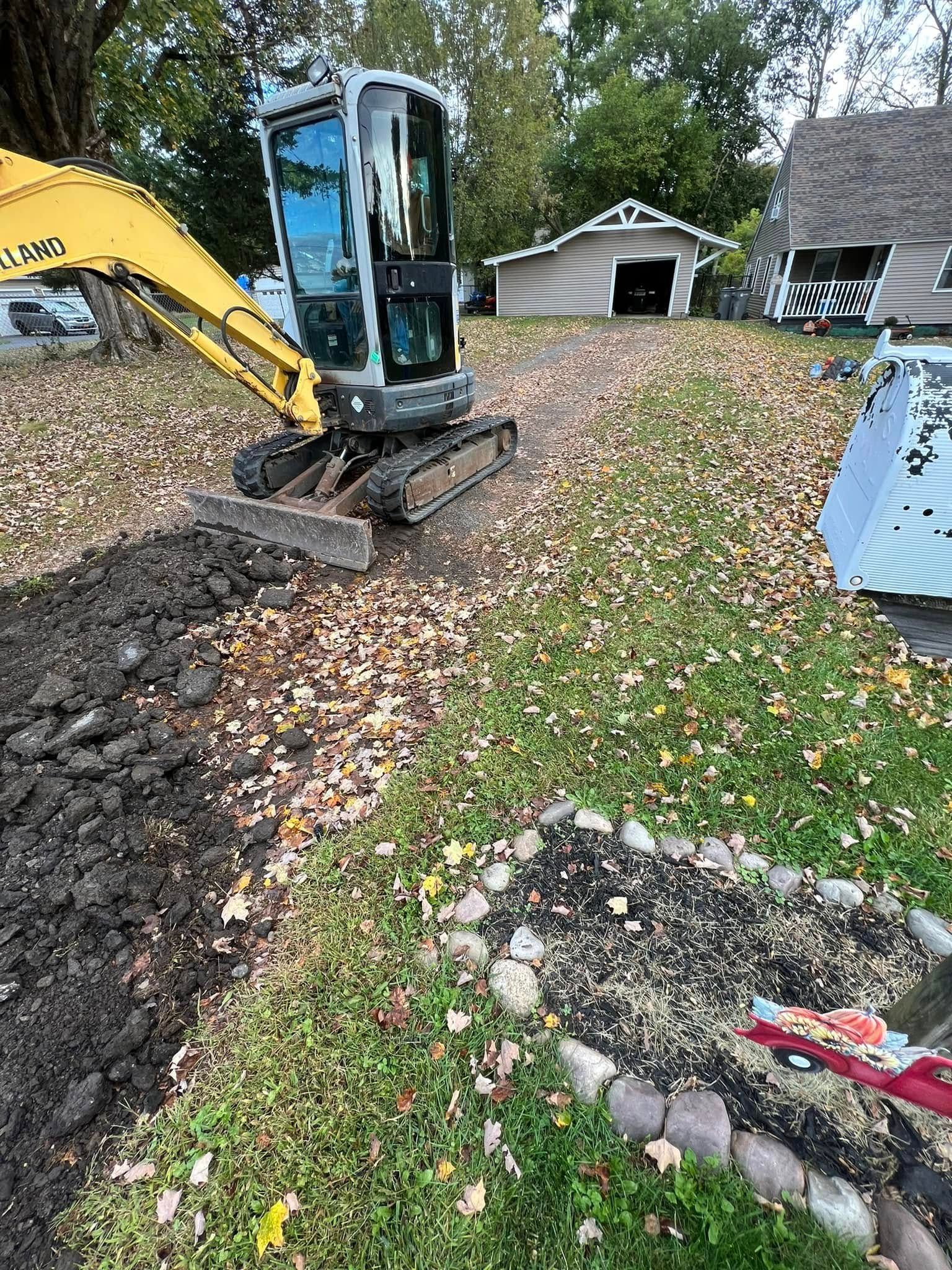Small excavator grading leaf-covered ground beside a house and garage in a yard