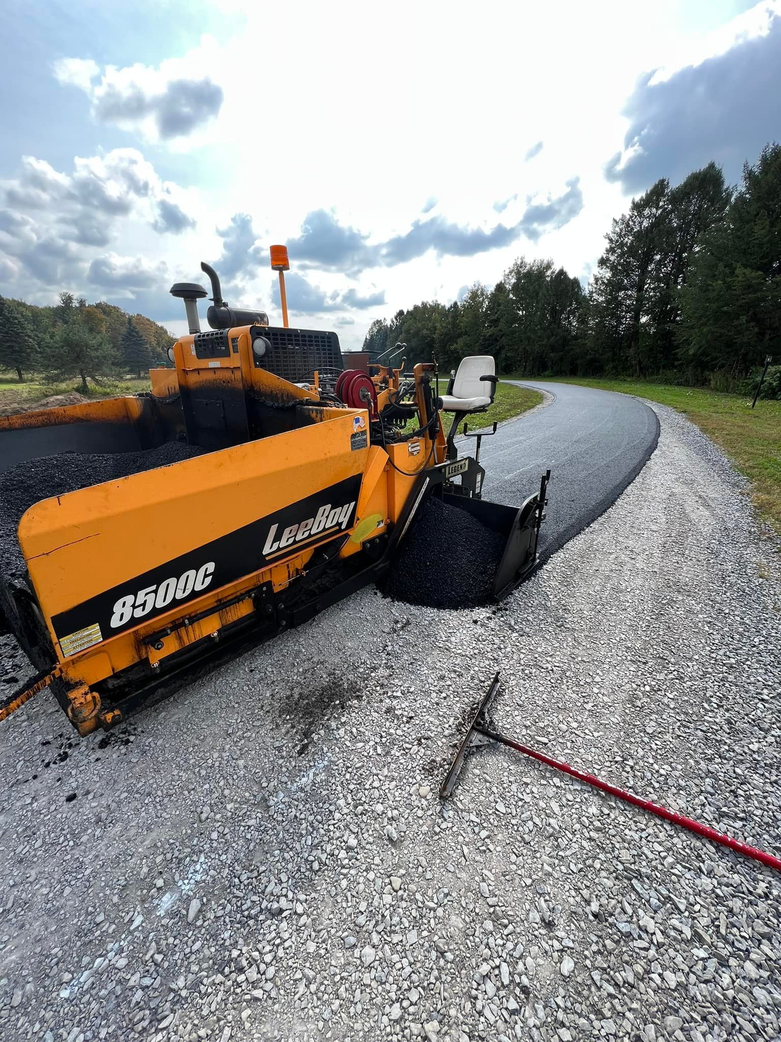 Orange asphalt paver laying gravel on a curved road under a cloudy sky