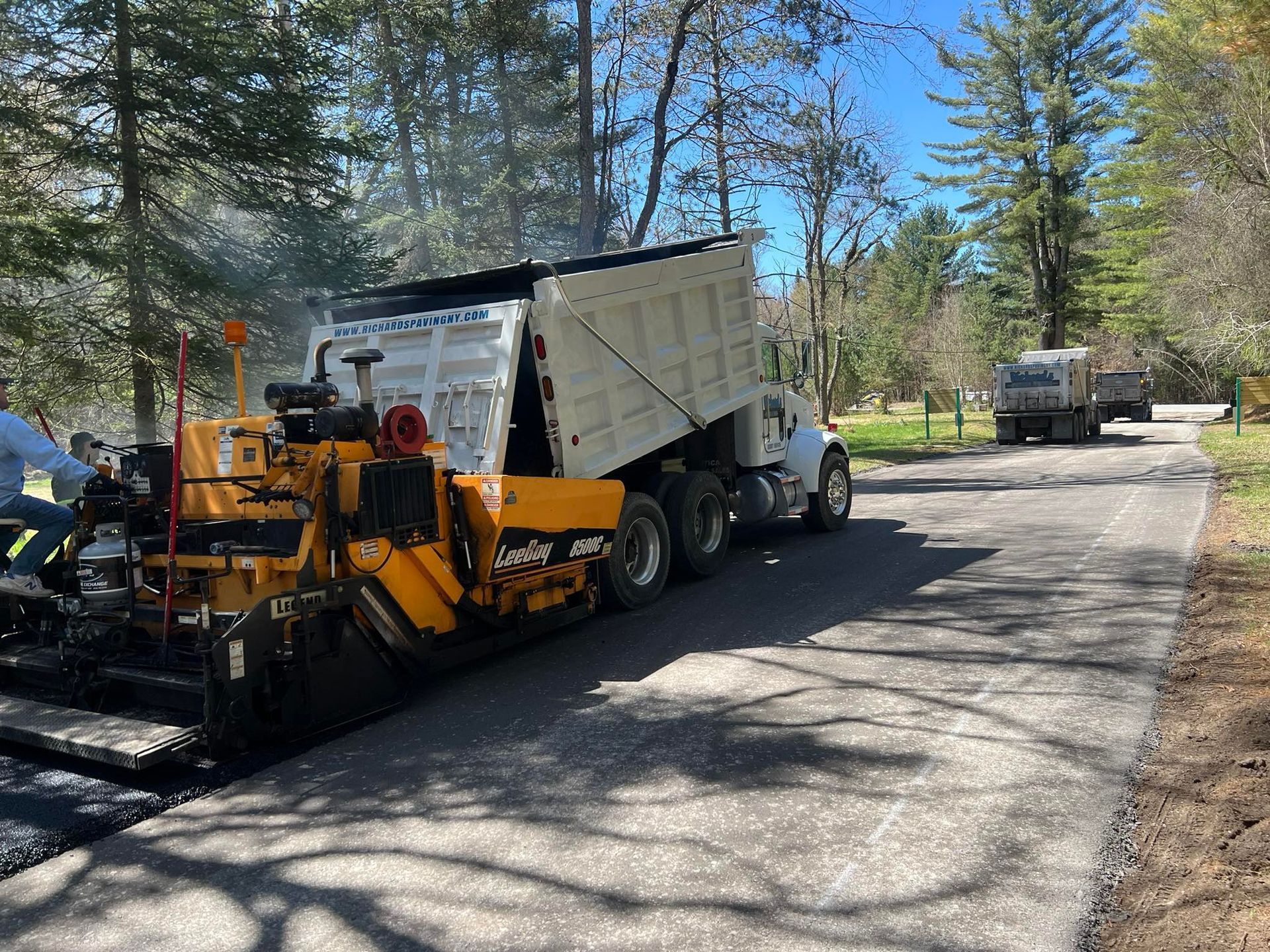Yellow paving machine and dump truck working on a tree-lined road, with another truck in the distance