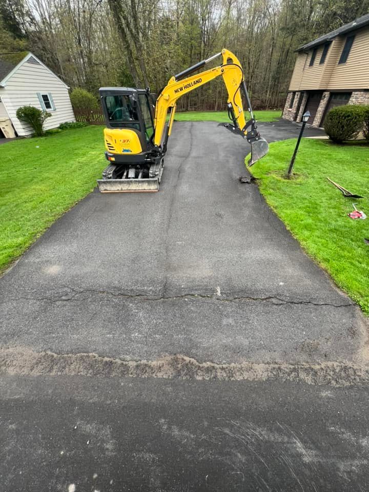 Small yellow excavator on a newly paved driveway between houses and green lawns
