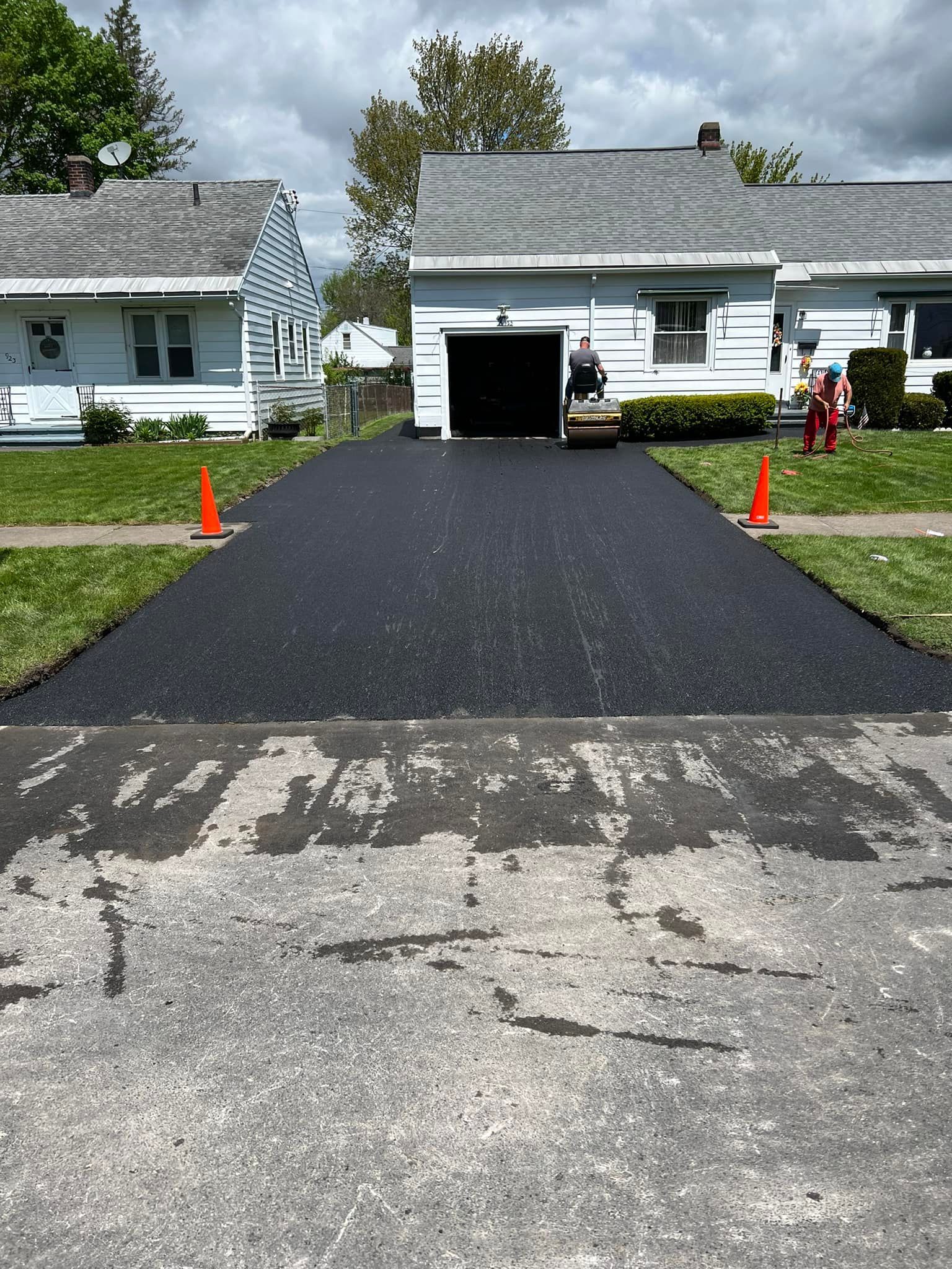 Fresh black asphalt driveway being installed between two white houses, with orange cones nearby.
