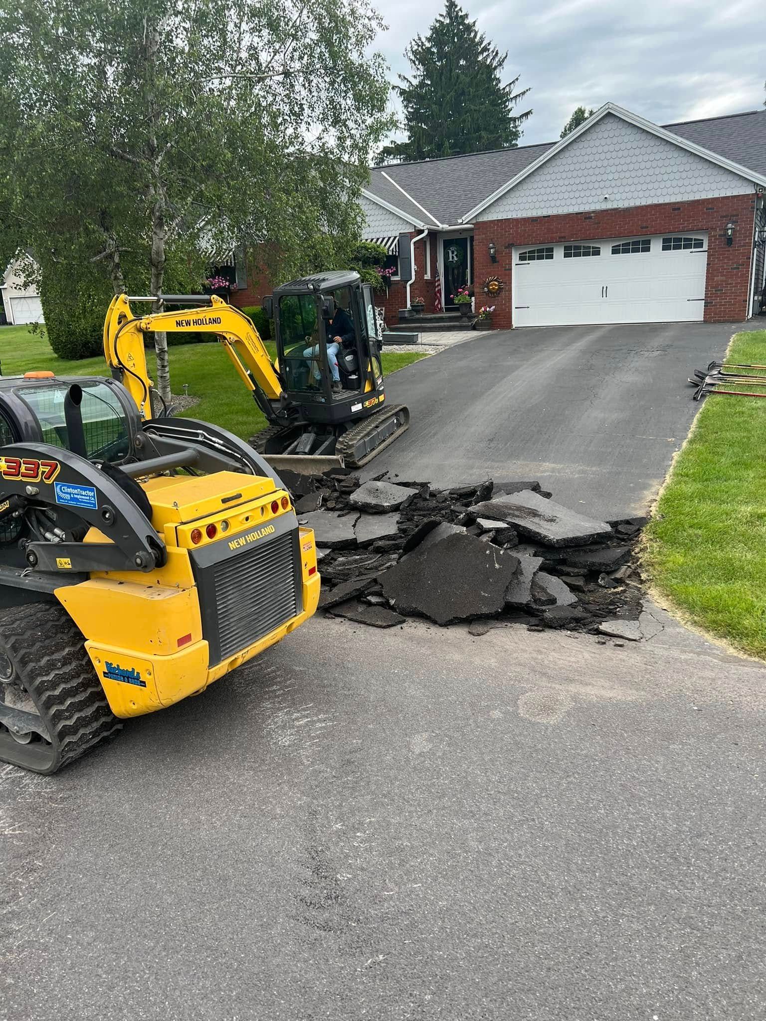 Yellow mini excavator repairing broken asphalt on a suburban driveway near a house with a garage.