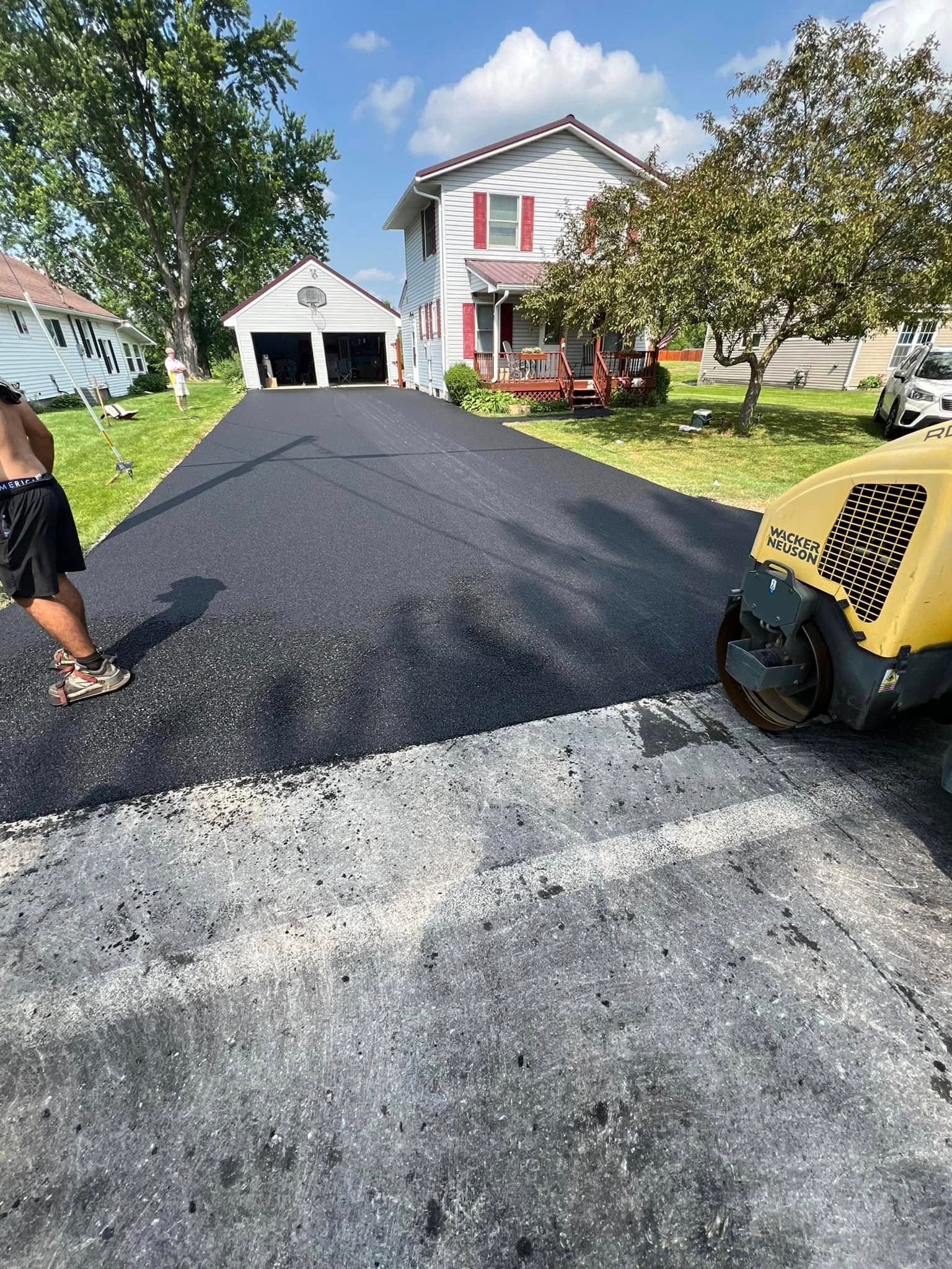 Fresh black asphalt driveway being paved outside a suburban house, with workers and a paving machine.