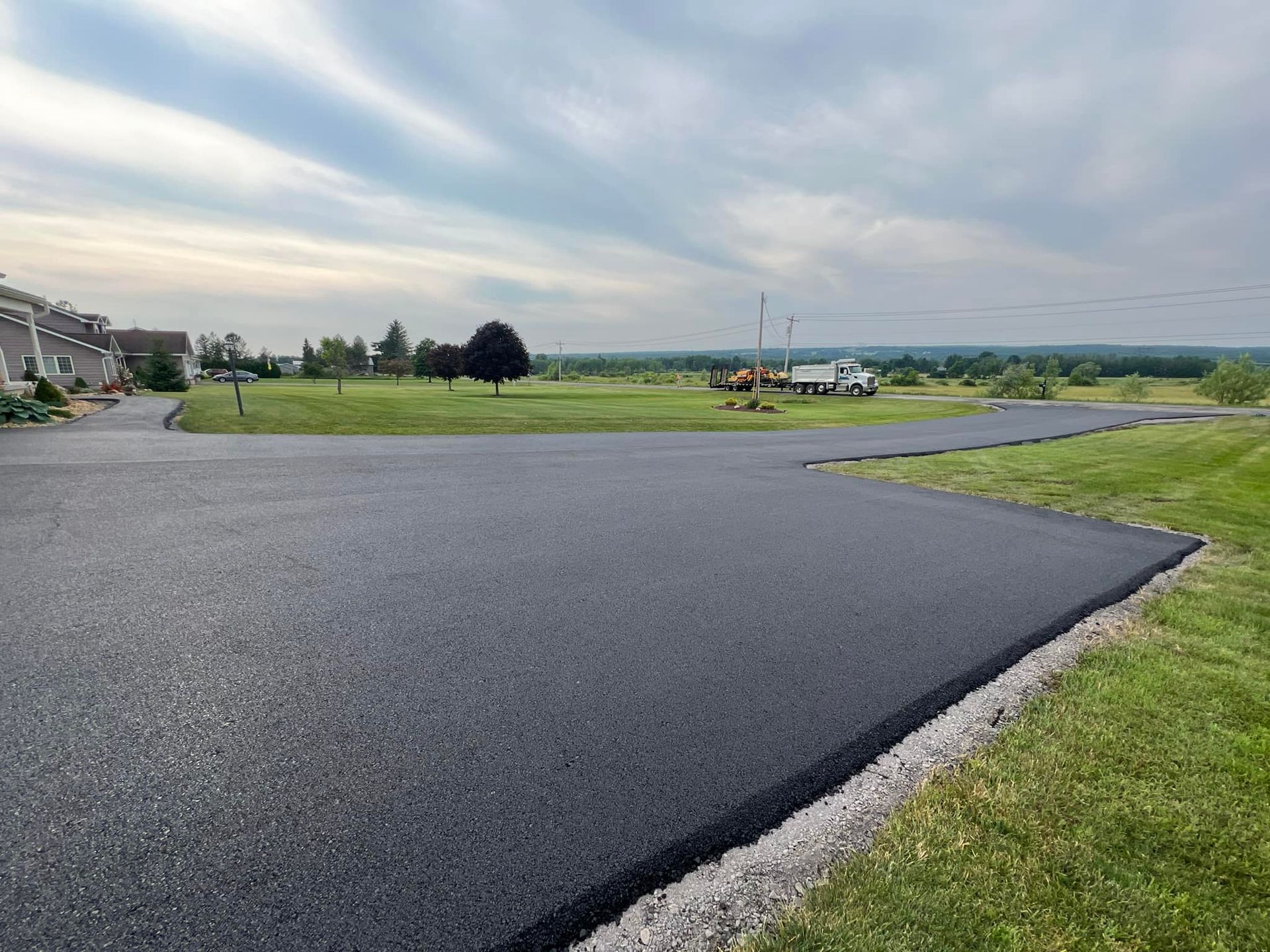 Freshly paved dark asphalt road in a grassy rural landscape under a cloudy sky