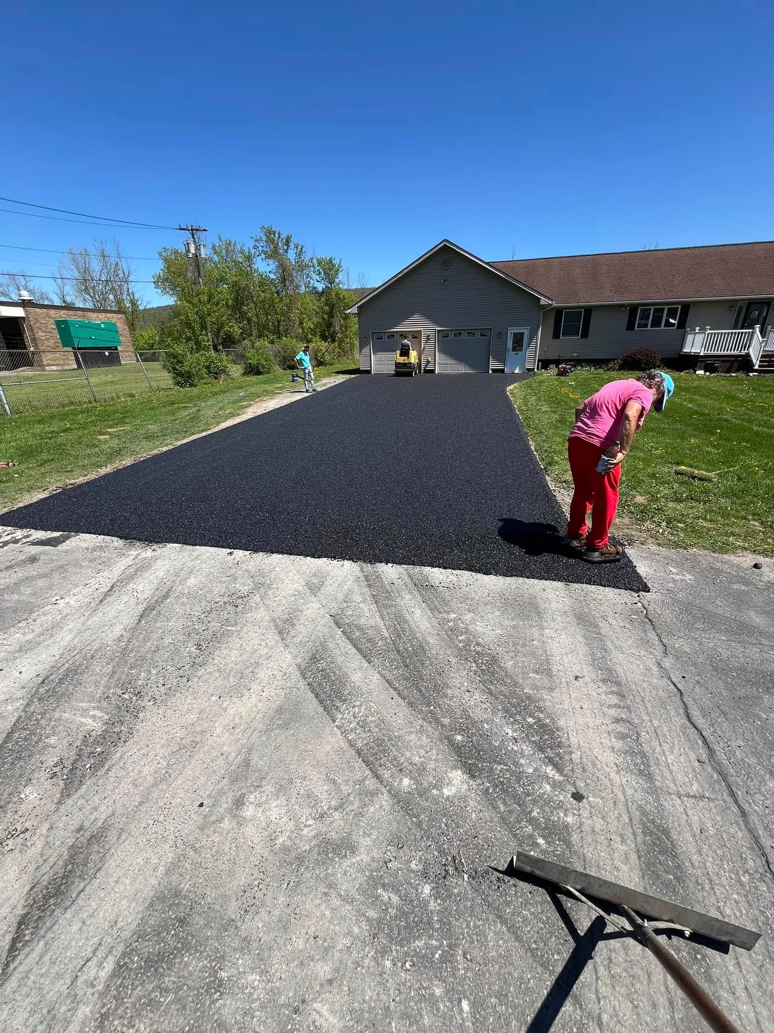 Worker spreading fresh black asphalt in a driveway beside a house on a sunny day