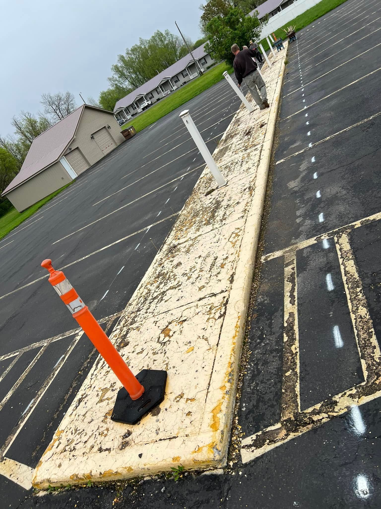 Tilted view of a parking lot with a long concrete barrier, orange cone, and a person walking near houses.