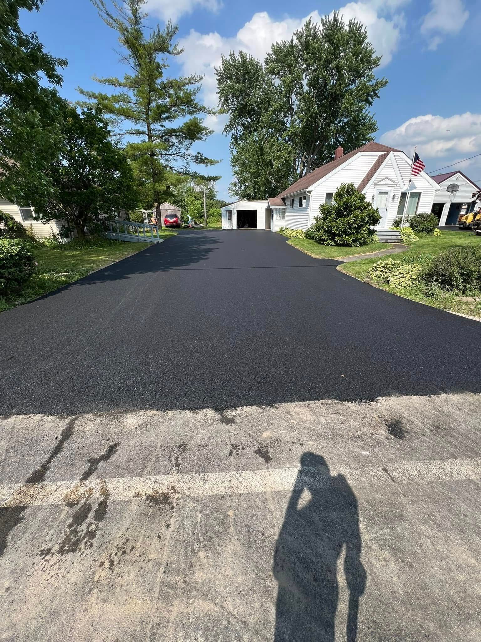 Freshly paved black driveway leading to a white house with trees and a blue sky