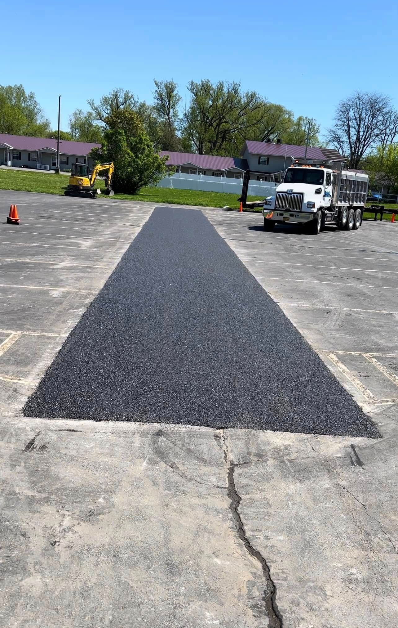 Freshly paved black asphalt strip on a concrete lot with a paving machine and cones nearby under a blue sky
