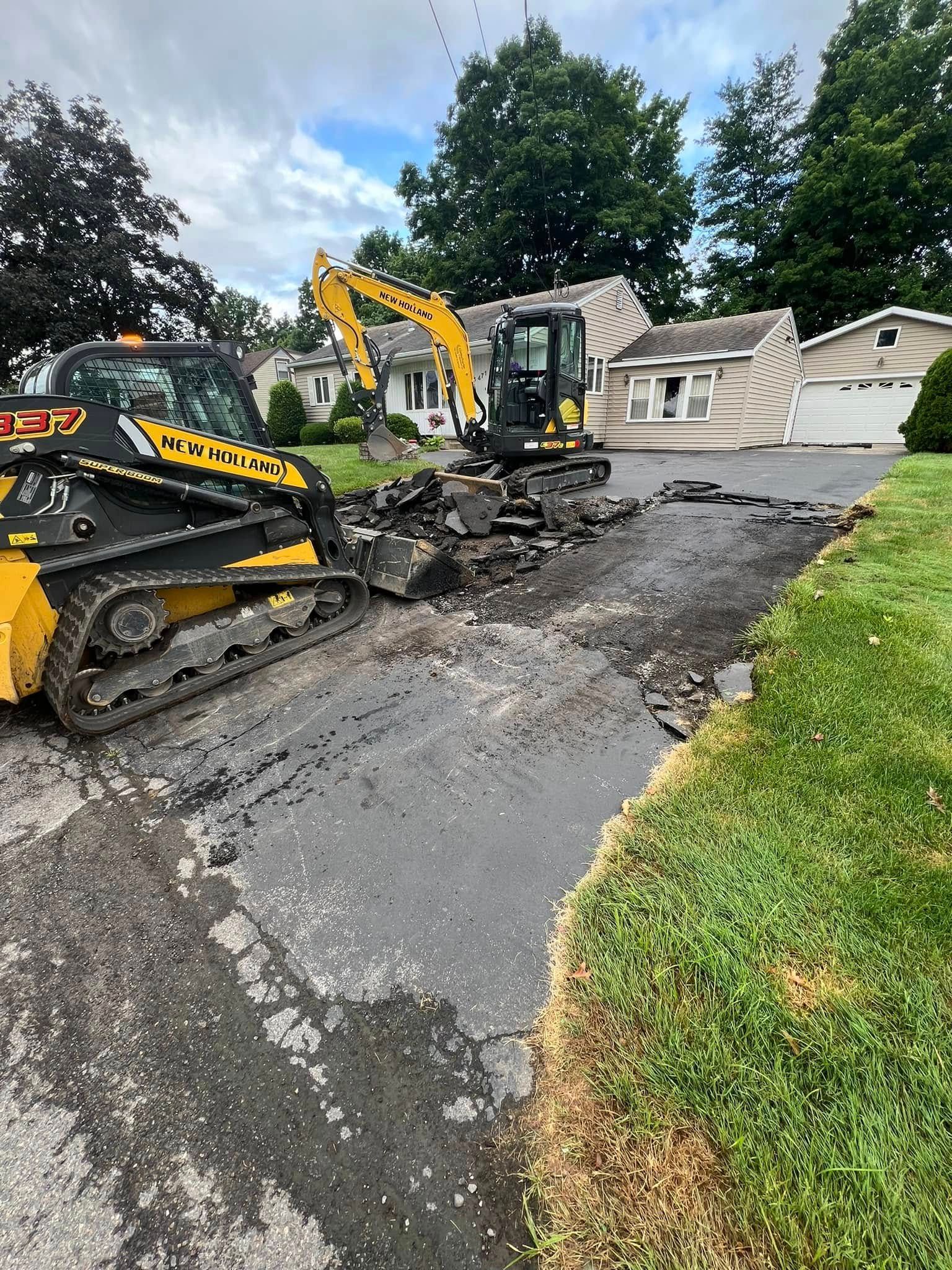 Two excavators tearing up a cracked driveway beside a white house and green lawn