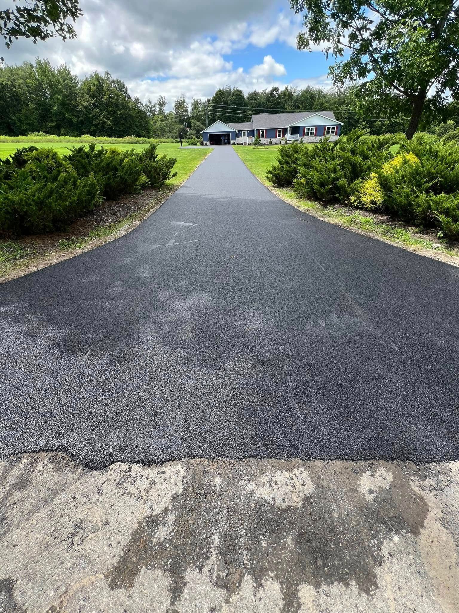 Gravel driveway leading to a white house, bordered by green hedges and trees under a bright sky