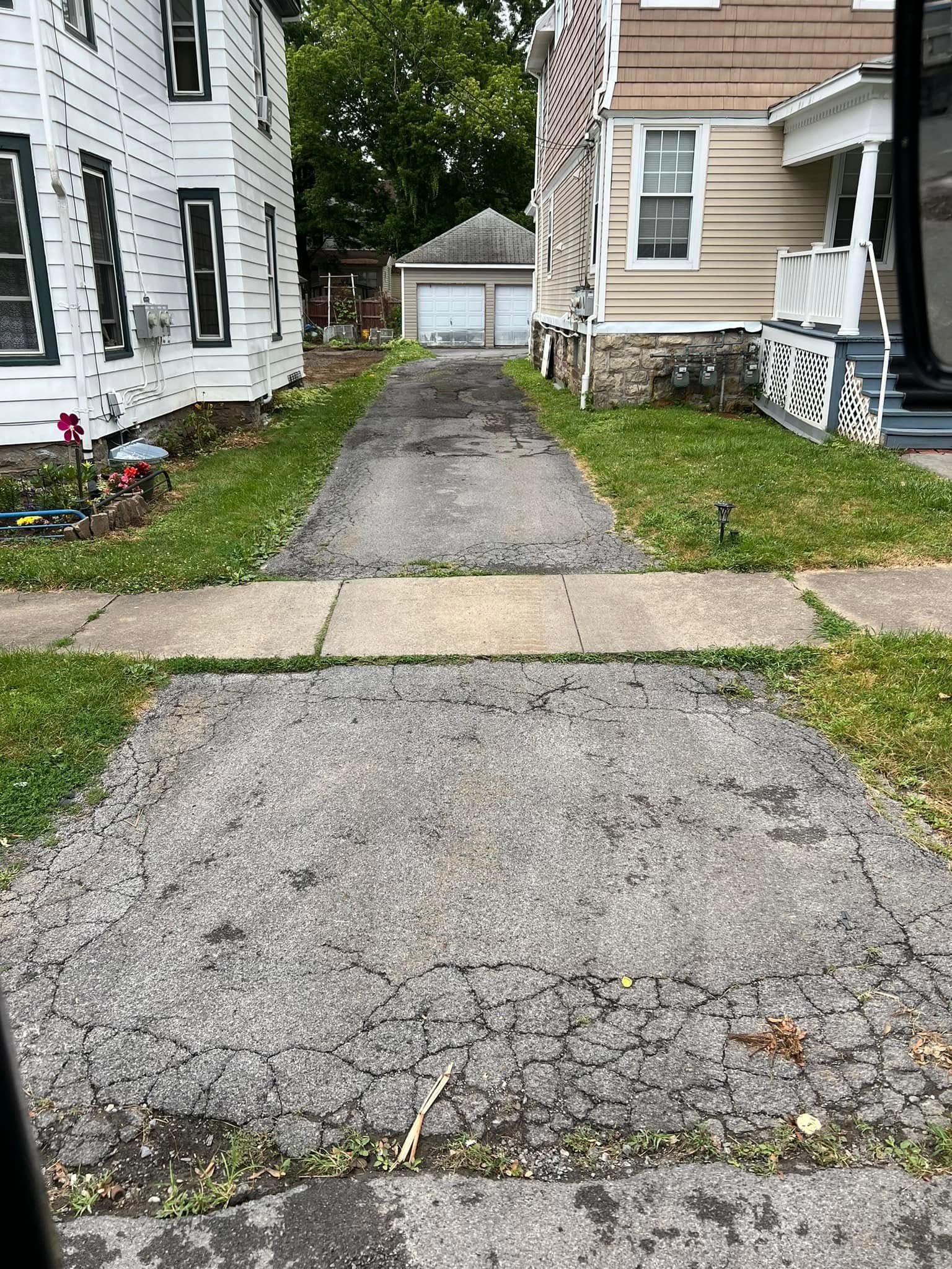 Gravel driveway between houses leading to a detached garage, bordered by grass and a sidewalk