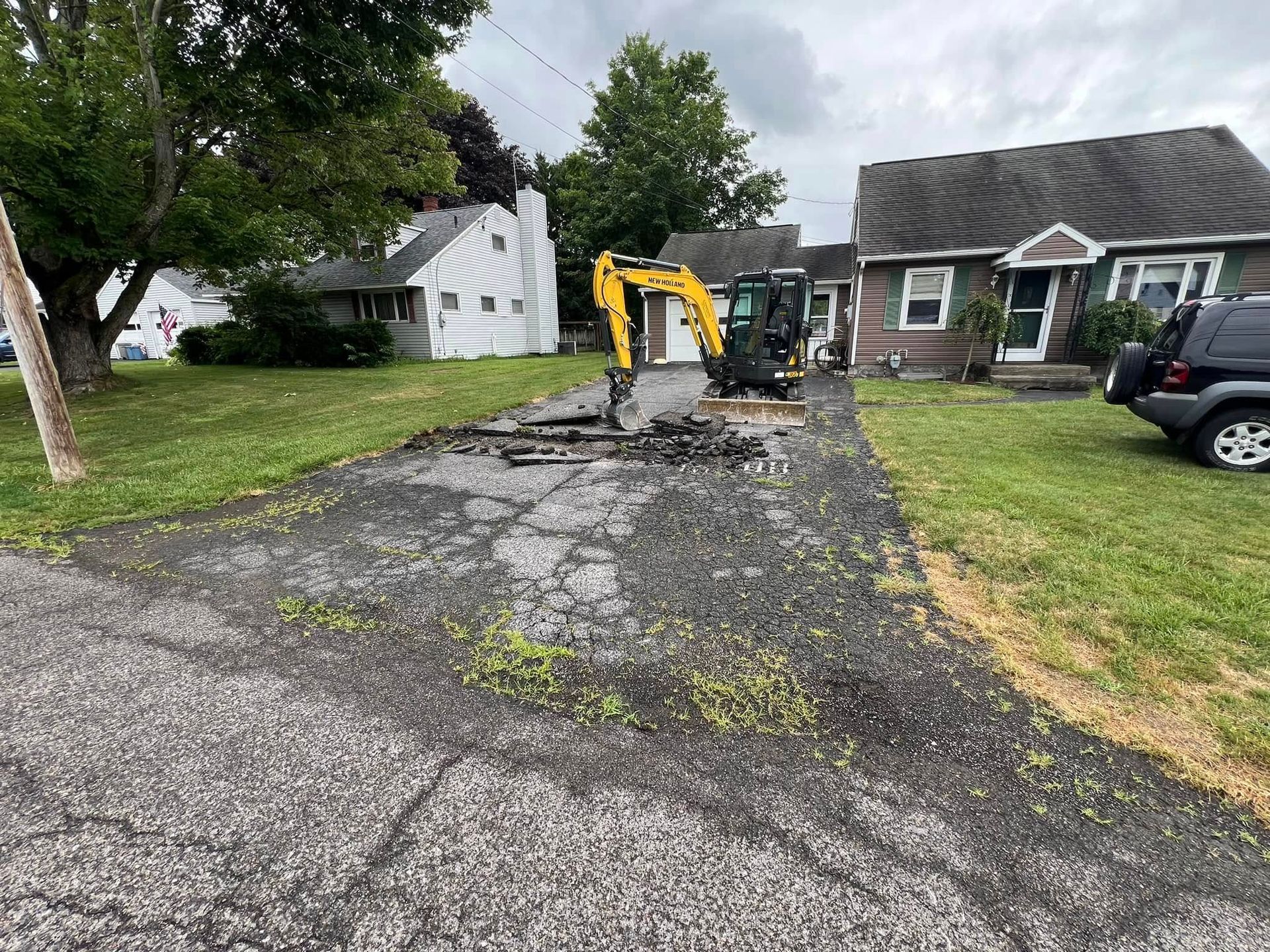 Yellow excavator repairing cracked driveway beside houses under overcast sky
