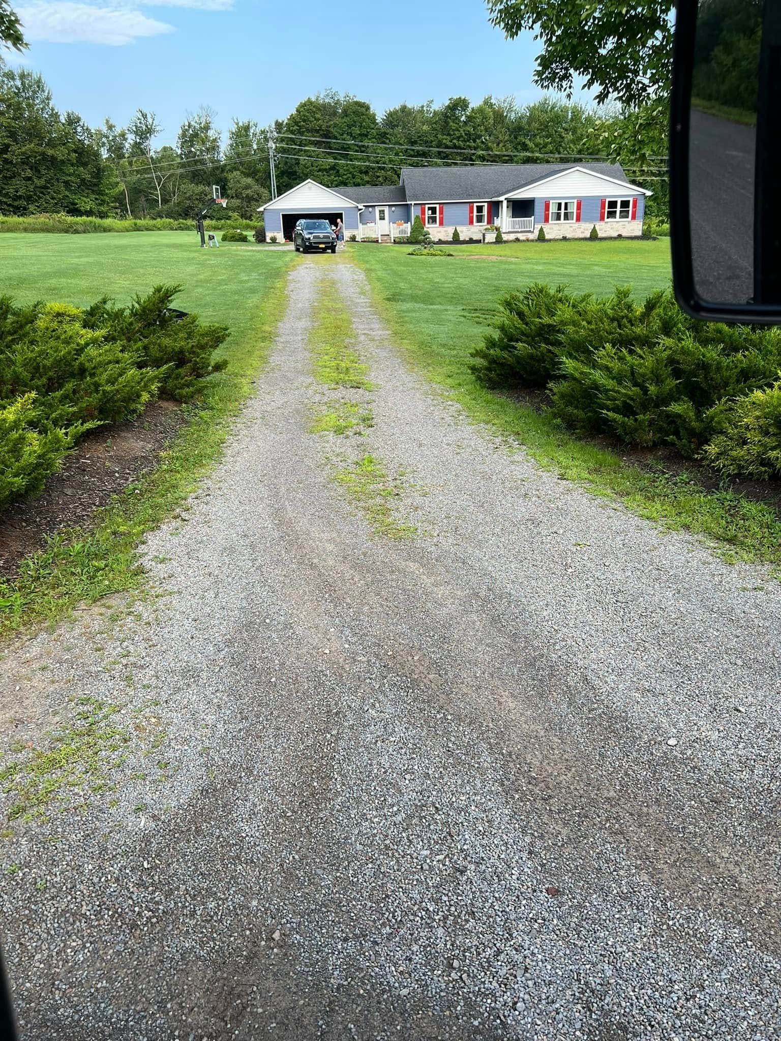 Gravel driveway leading to a white house with a red roof, framed by green lawn and trees.
