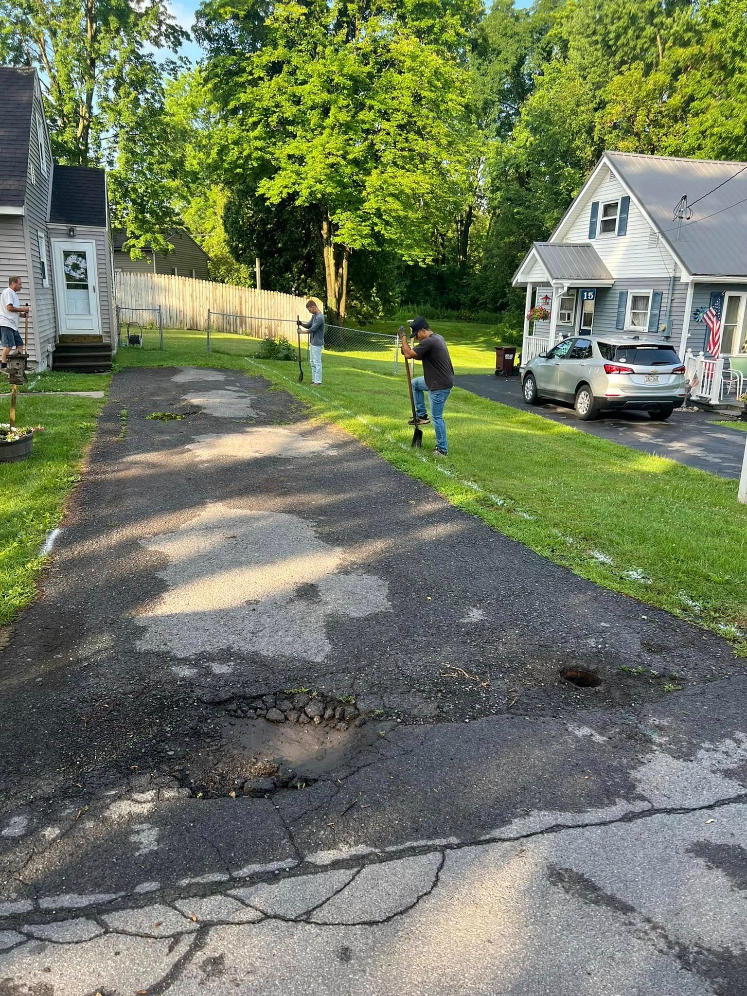 Damaged gravel driveway with potholes beside homes; a person stands near a parked car in the yard.