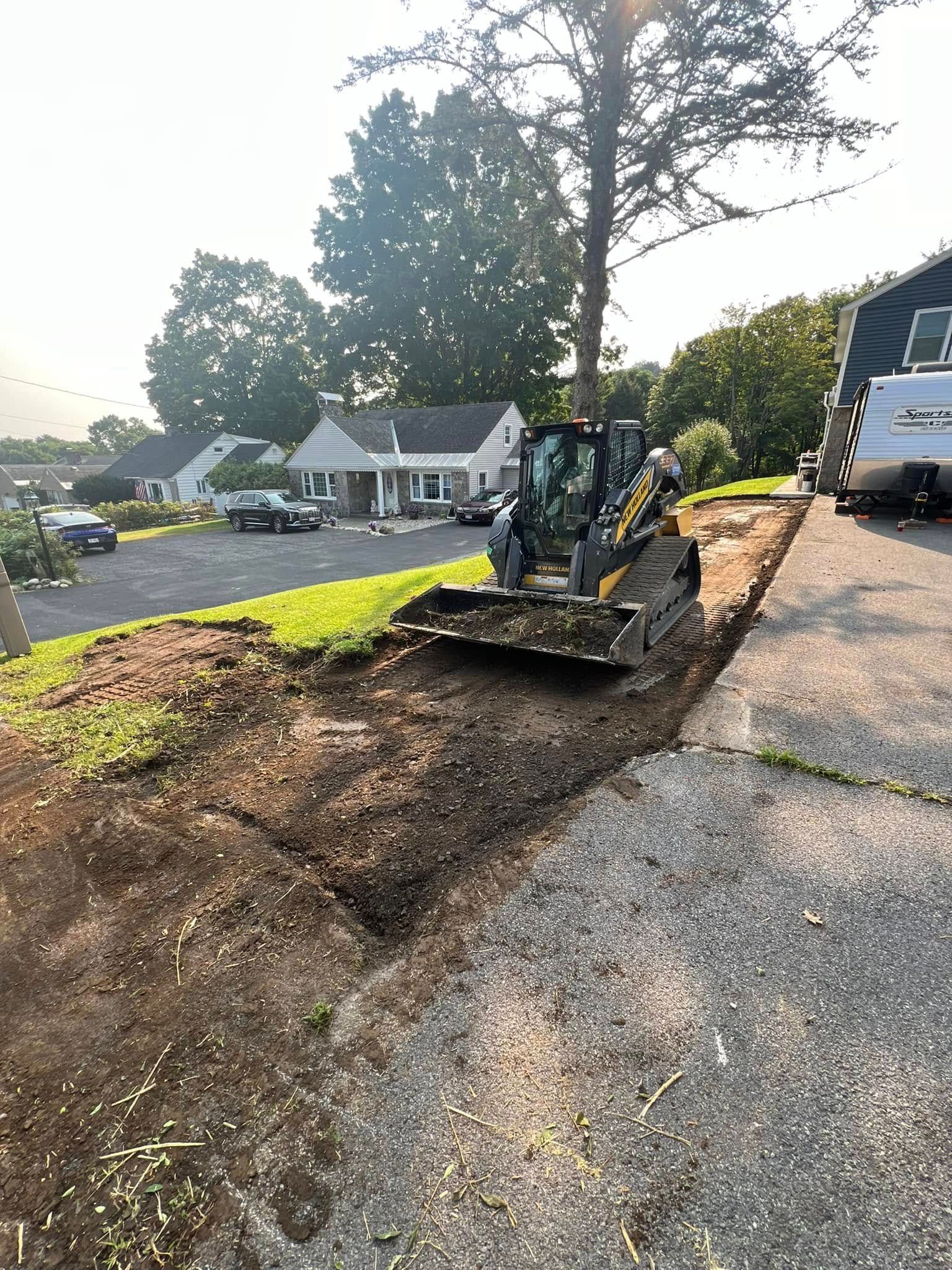 Backhoe clearing dirt along a roadside beside parked trucks and houses