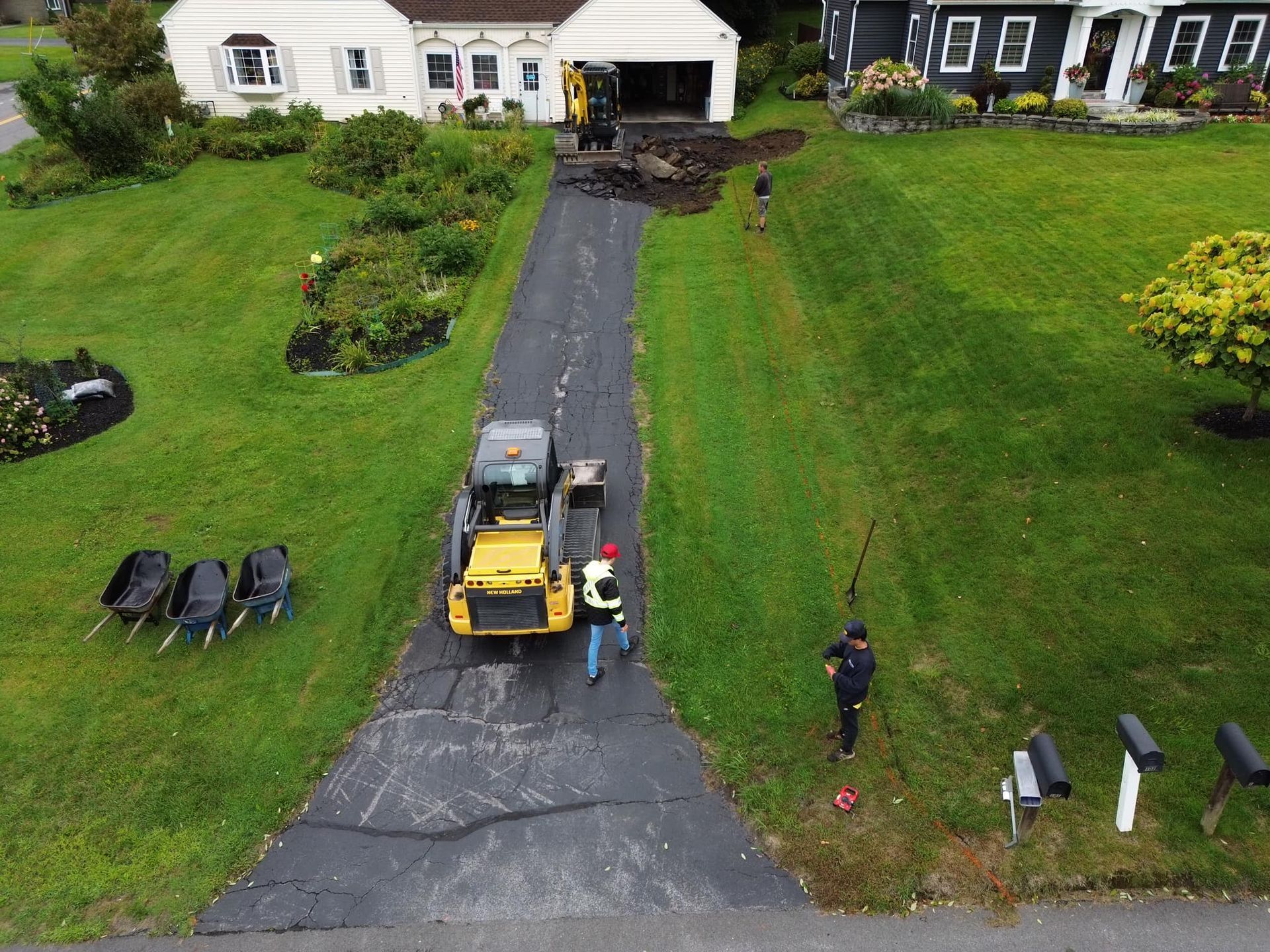Yellow tractor on a black driveway beside a white house, with two people and a green lawn around it.
