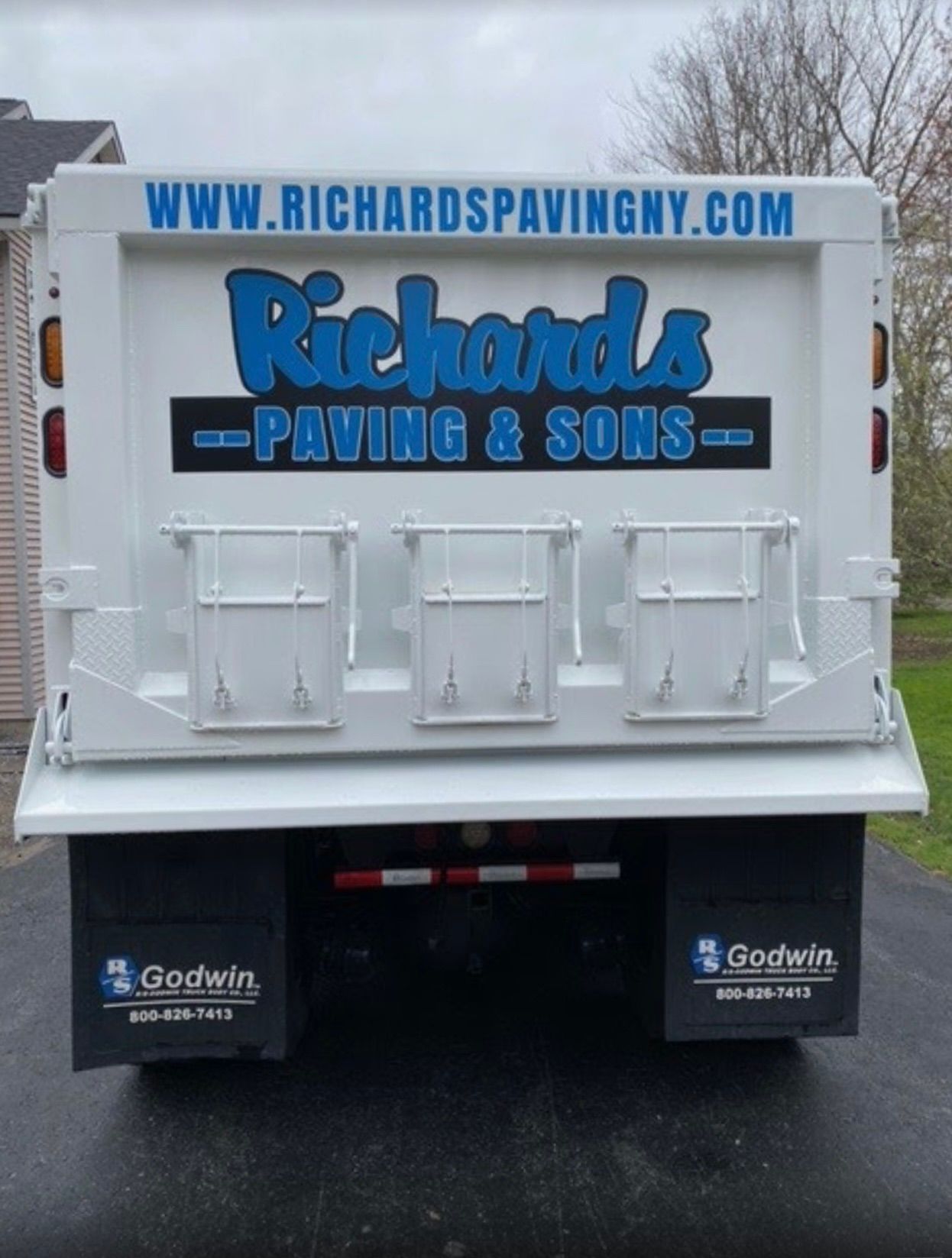 White dump truck tailgate with blue “Richards Paving & Sons” signage and website on a wet street.