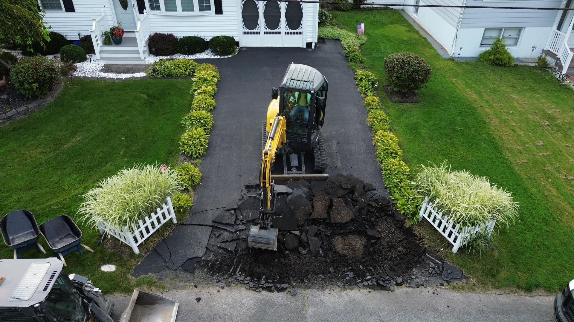 Excavator digging up a front driveway beside a white house and green lawn