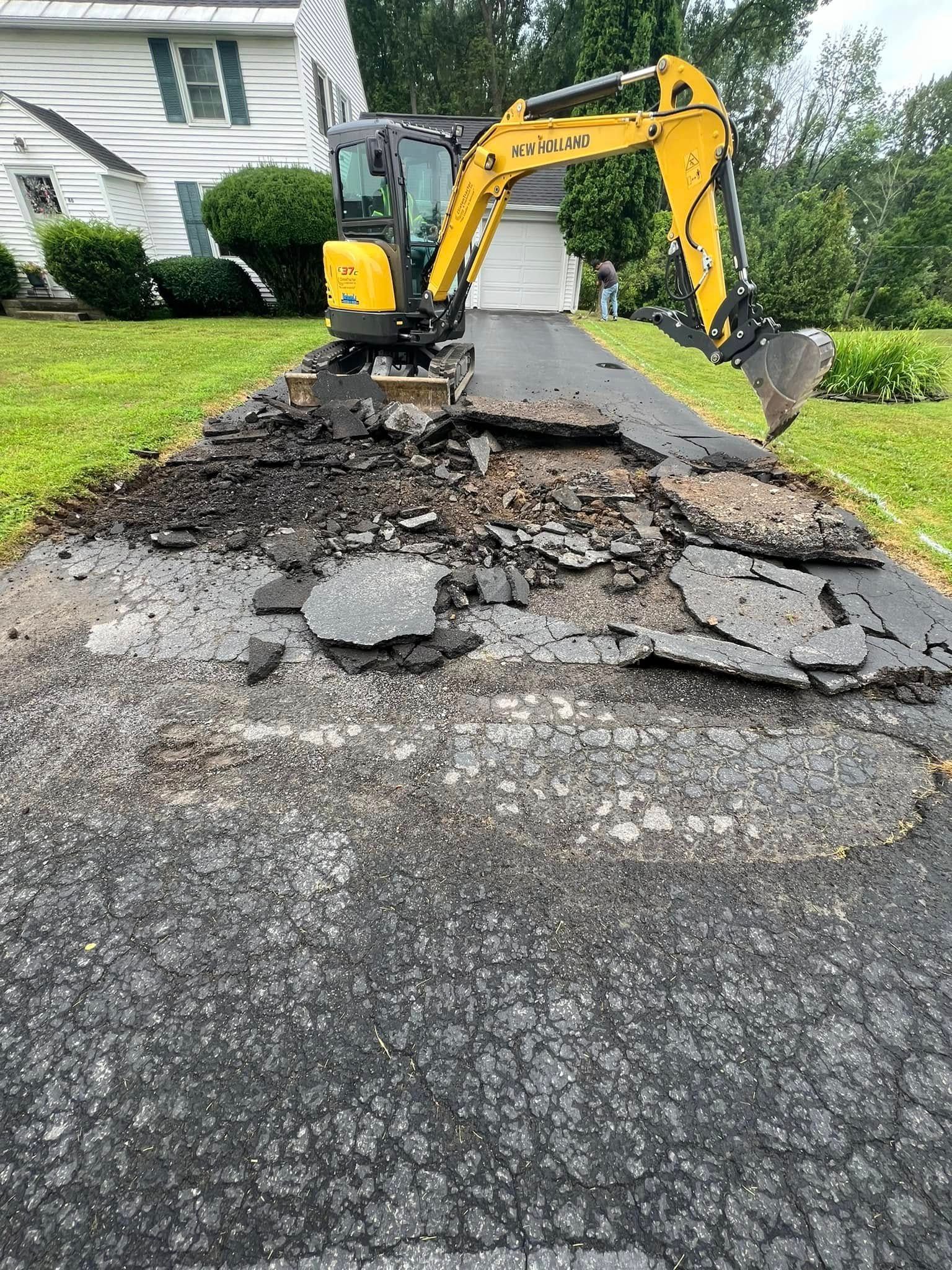 Yellow excavator tearing up cracked asphalt in a residential driveway, with house and lawn nearby