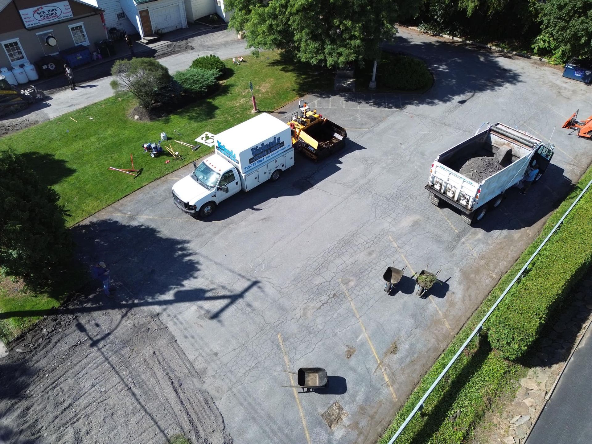 Aerial view of a work truck, dump truck, and equipment on a gravel lot beside a grassy area.