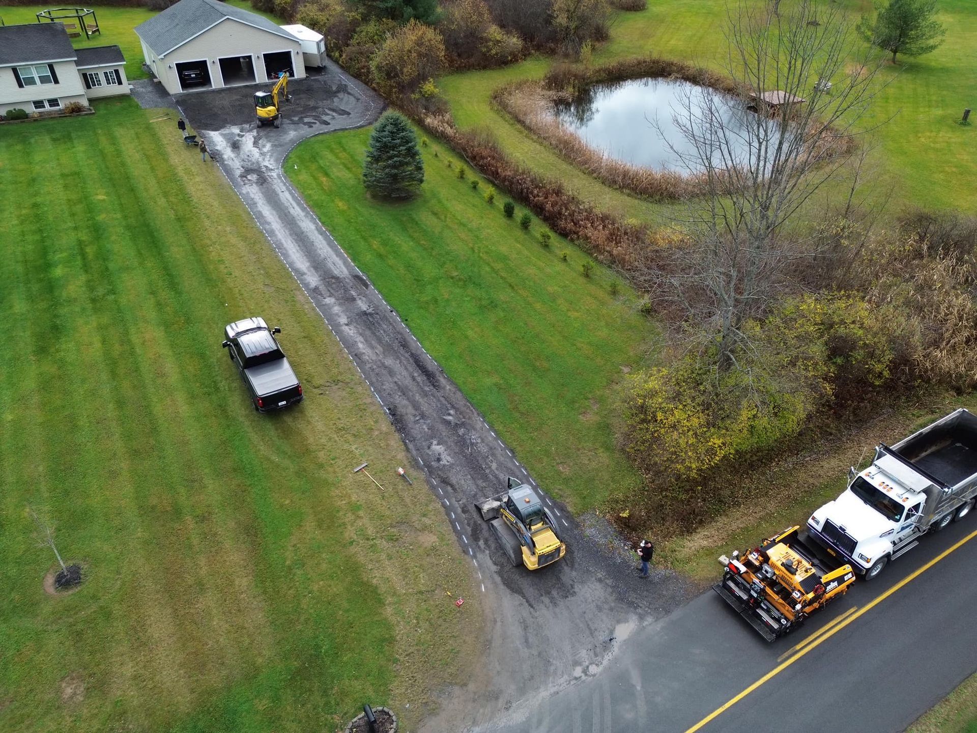Aerial view of a driveway by a house, with vehicles and a yellow paving machine on a rural property.