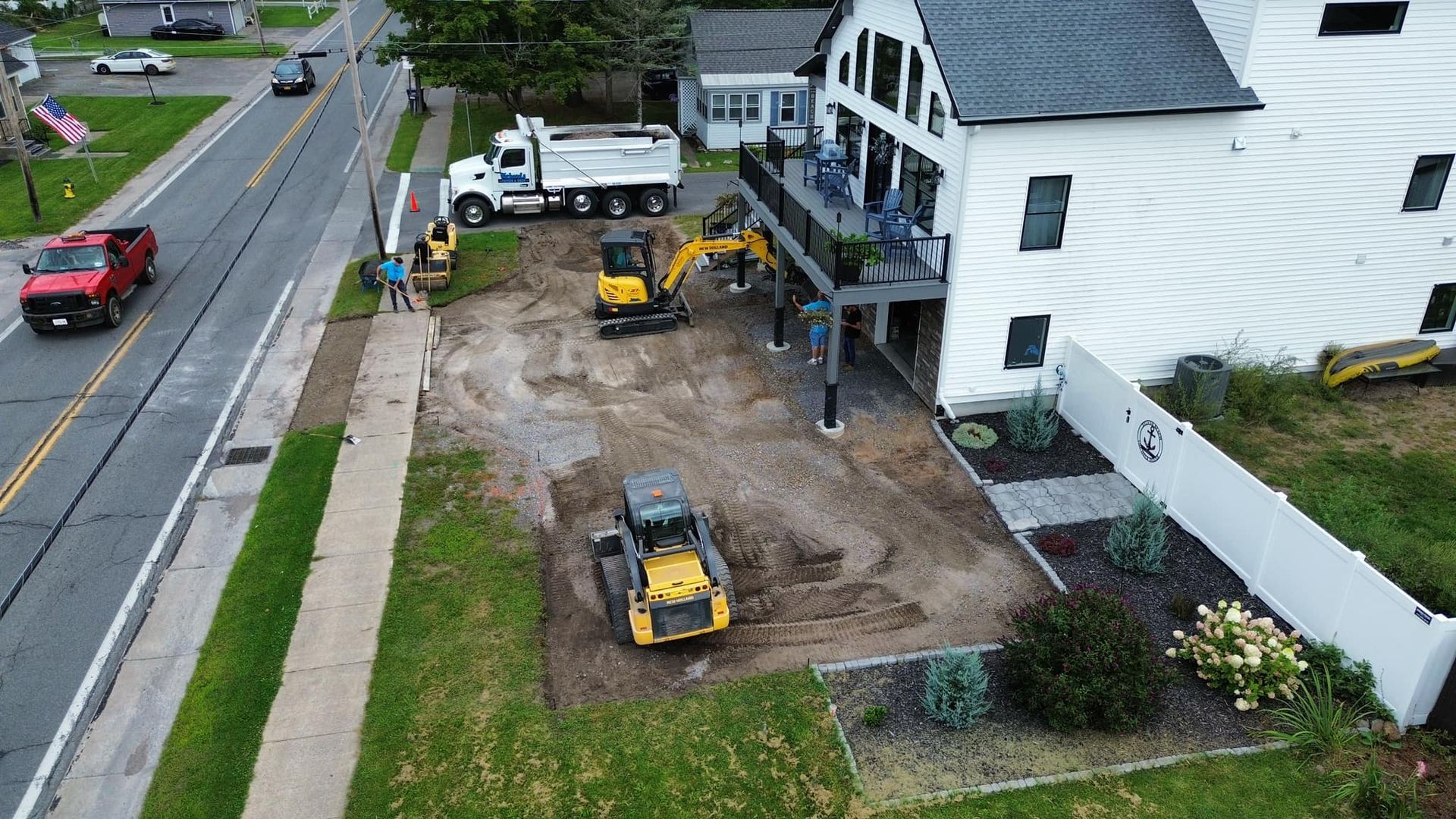 Construction equipment working in a small front yard beside a white house near a roadside