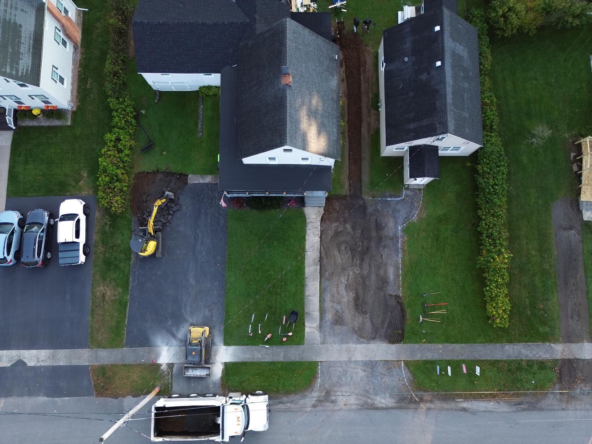 Aerial view of a residential street with houses, driveways, lawns, parked vehicles, and a sidewalk.