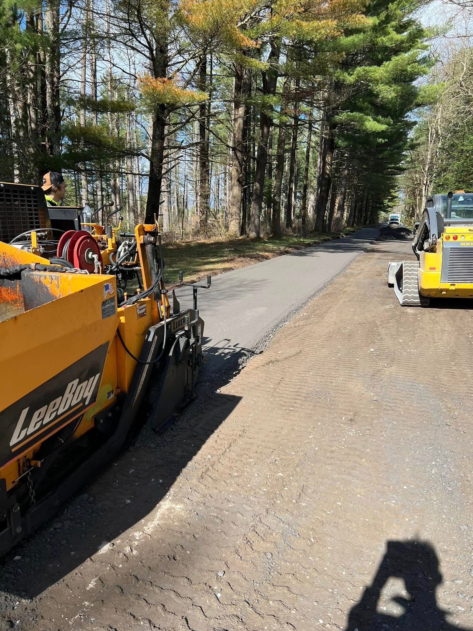 Roadwork scene with yellow paving machines on a tree-lined road and a worker in a safety vest.