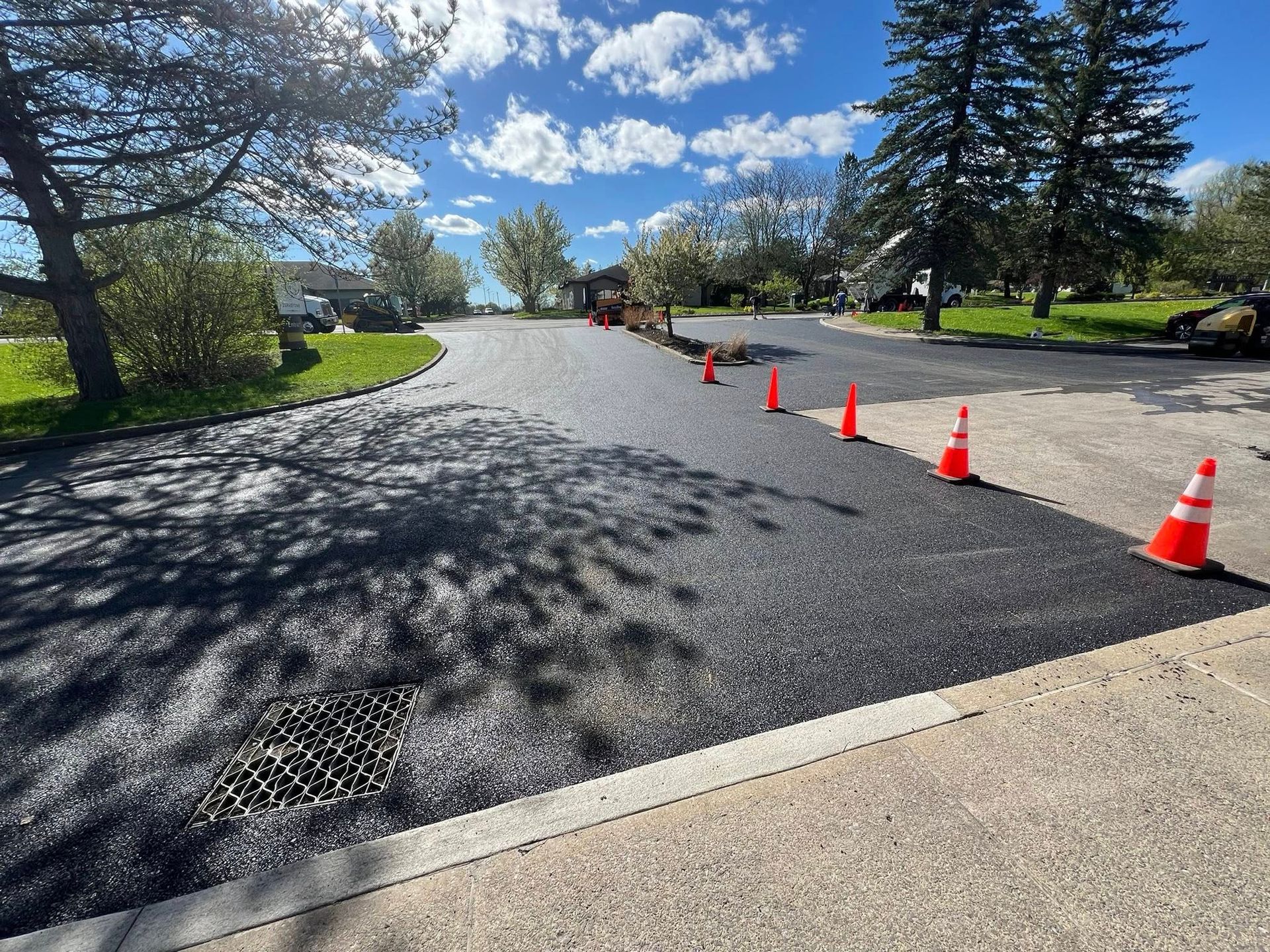 Freshly paved road curving through a park, marked by orange traffic cones under a bright blue sky.