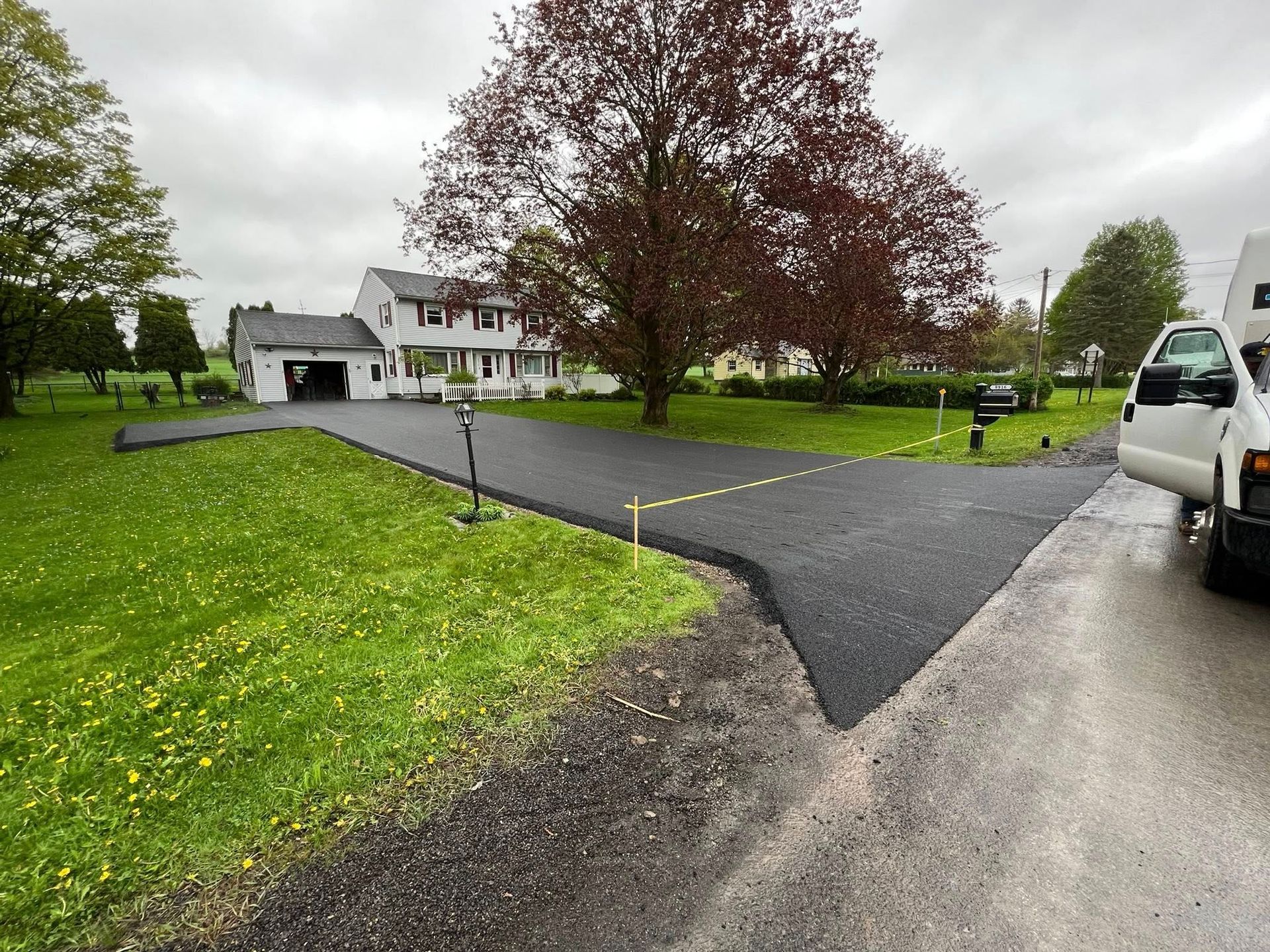 Driveway curving through a green yard toward a house, with a white truck parked at right under cloudy skies.