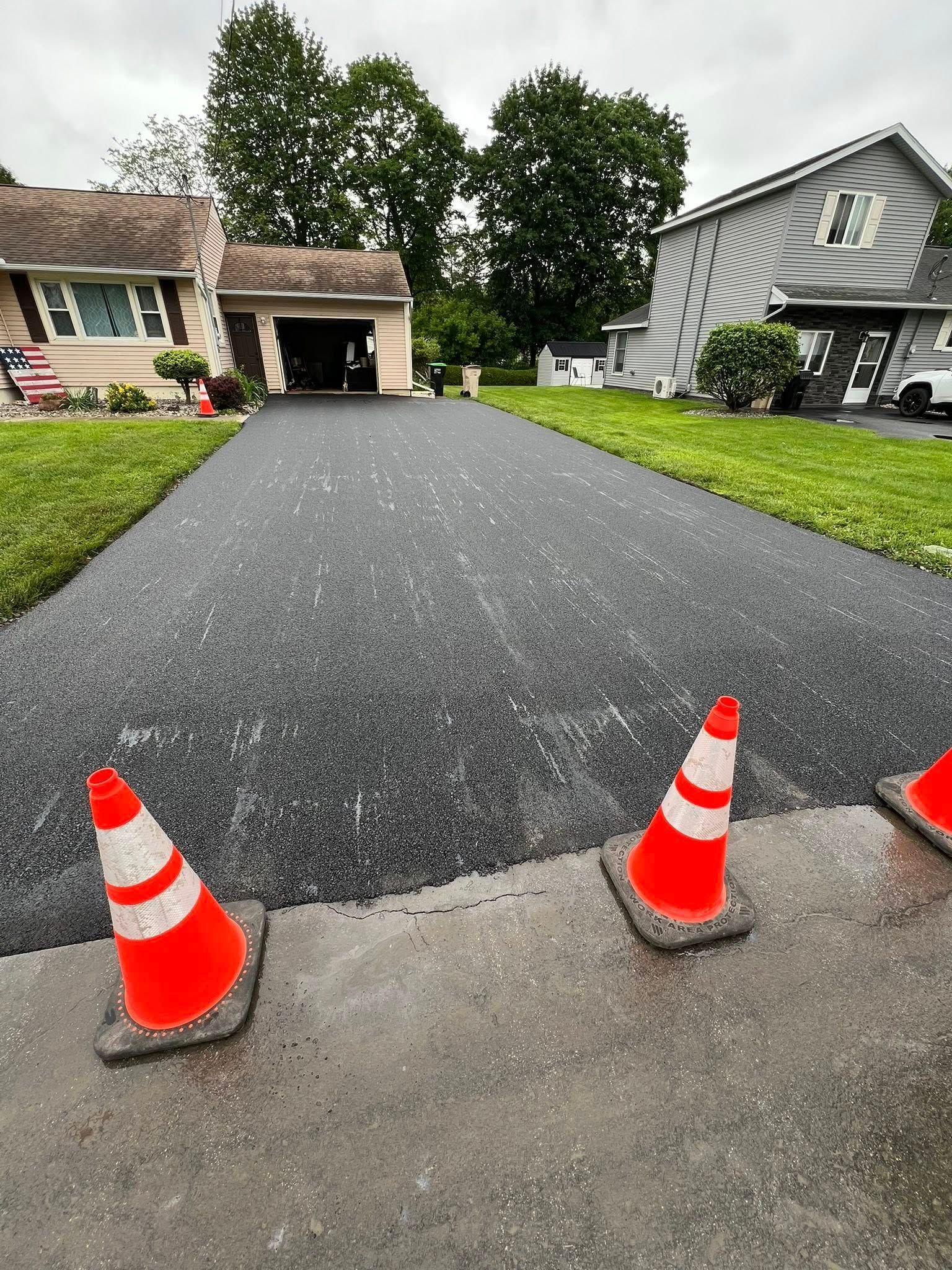 Fresh blacktop driveway with orange traffic cones at the entrance, suburban houses in the background