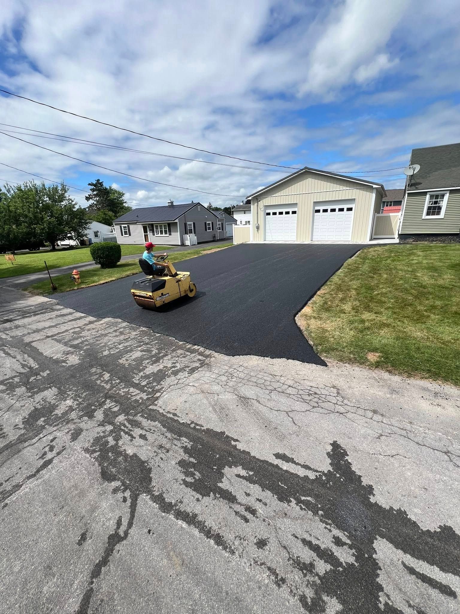Boy riding a yellow toy bulldozer on a driveway in front of a garage and house