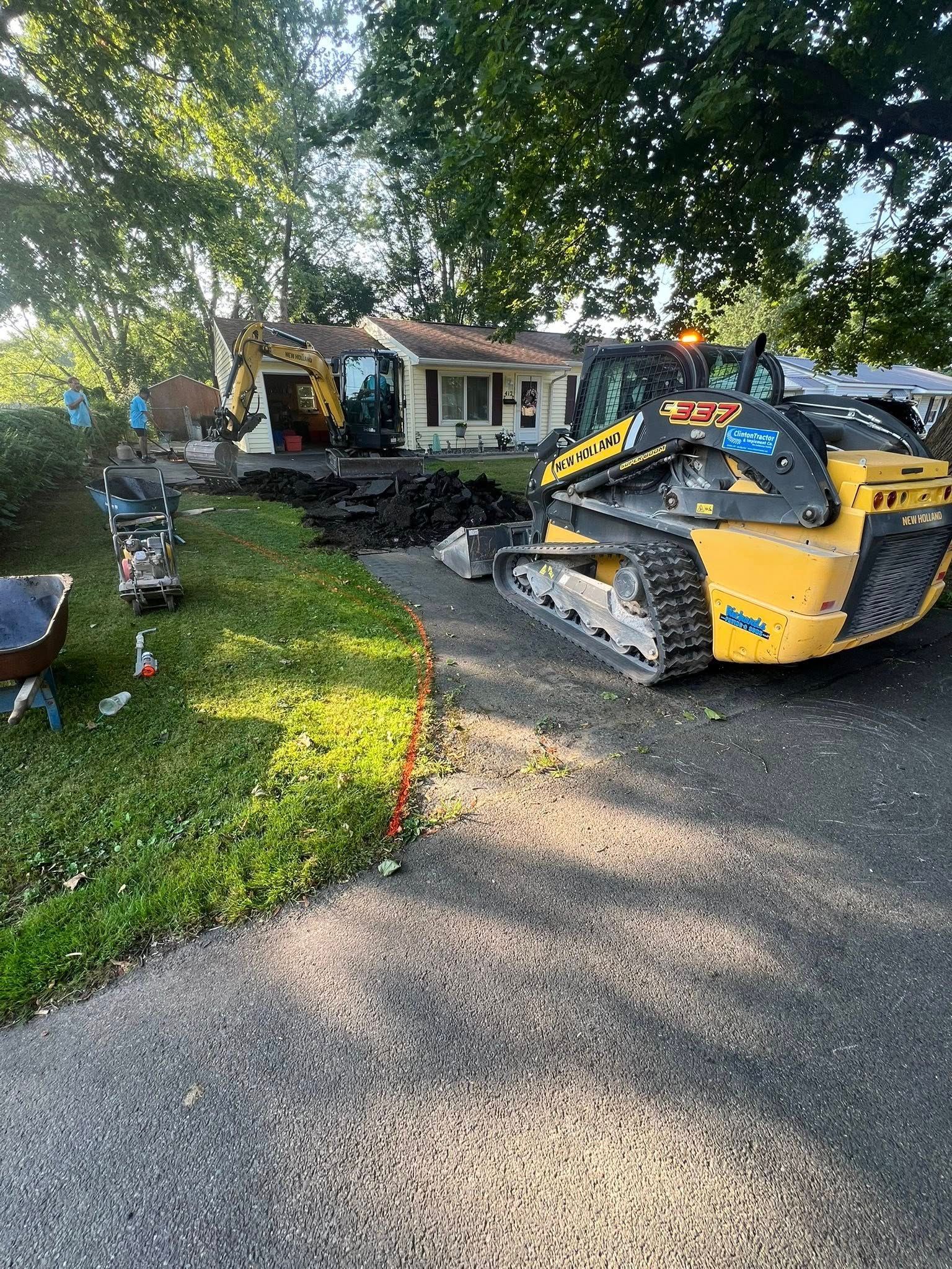 Yellow construction vehicle in a front yard beside a house, with debris and a driveway under trees.