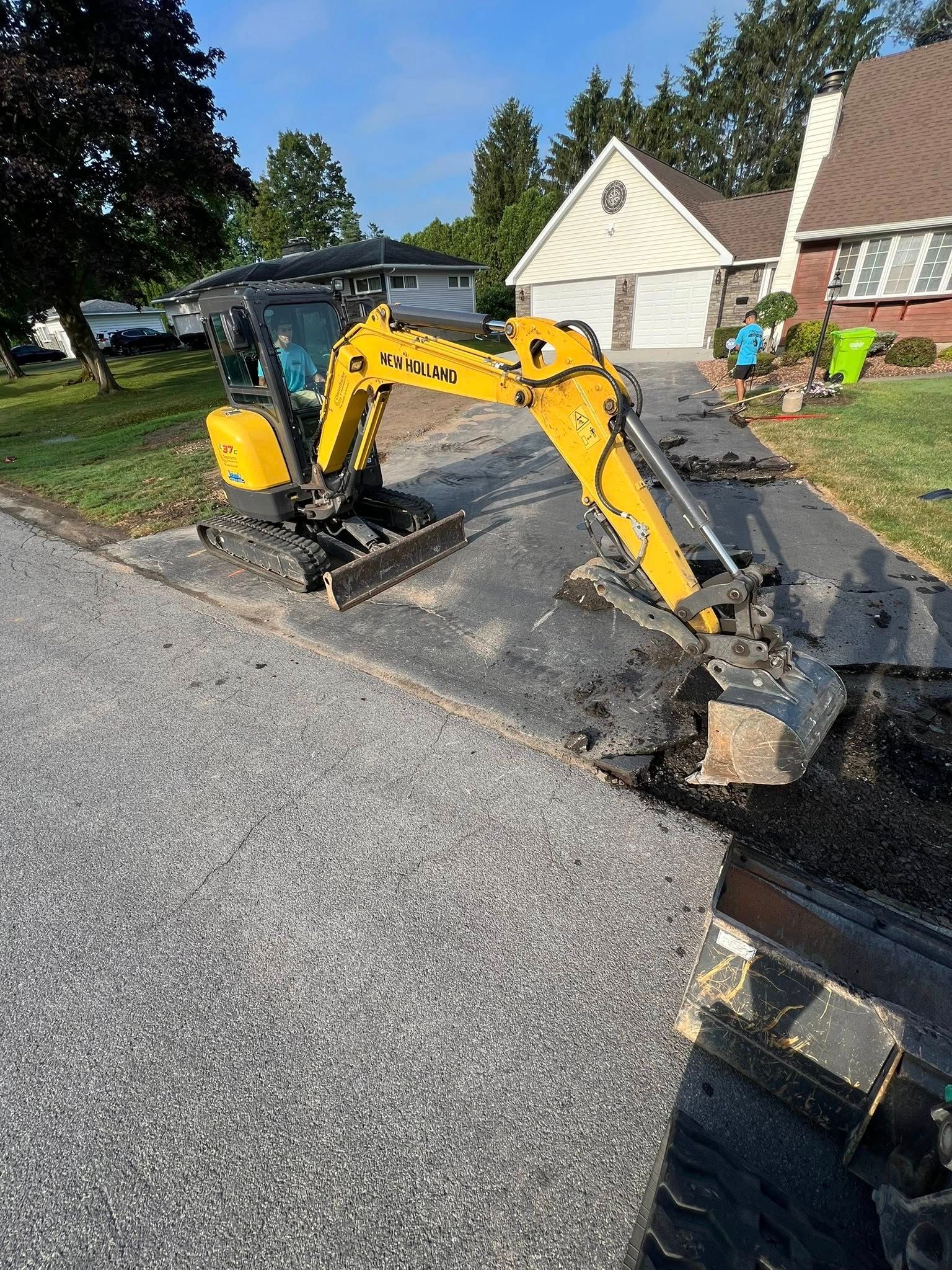 Yellow excavator digging a trench beside a paved road in a suburban driveway area