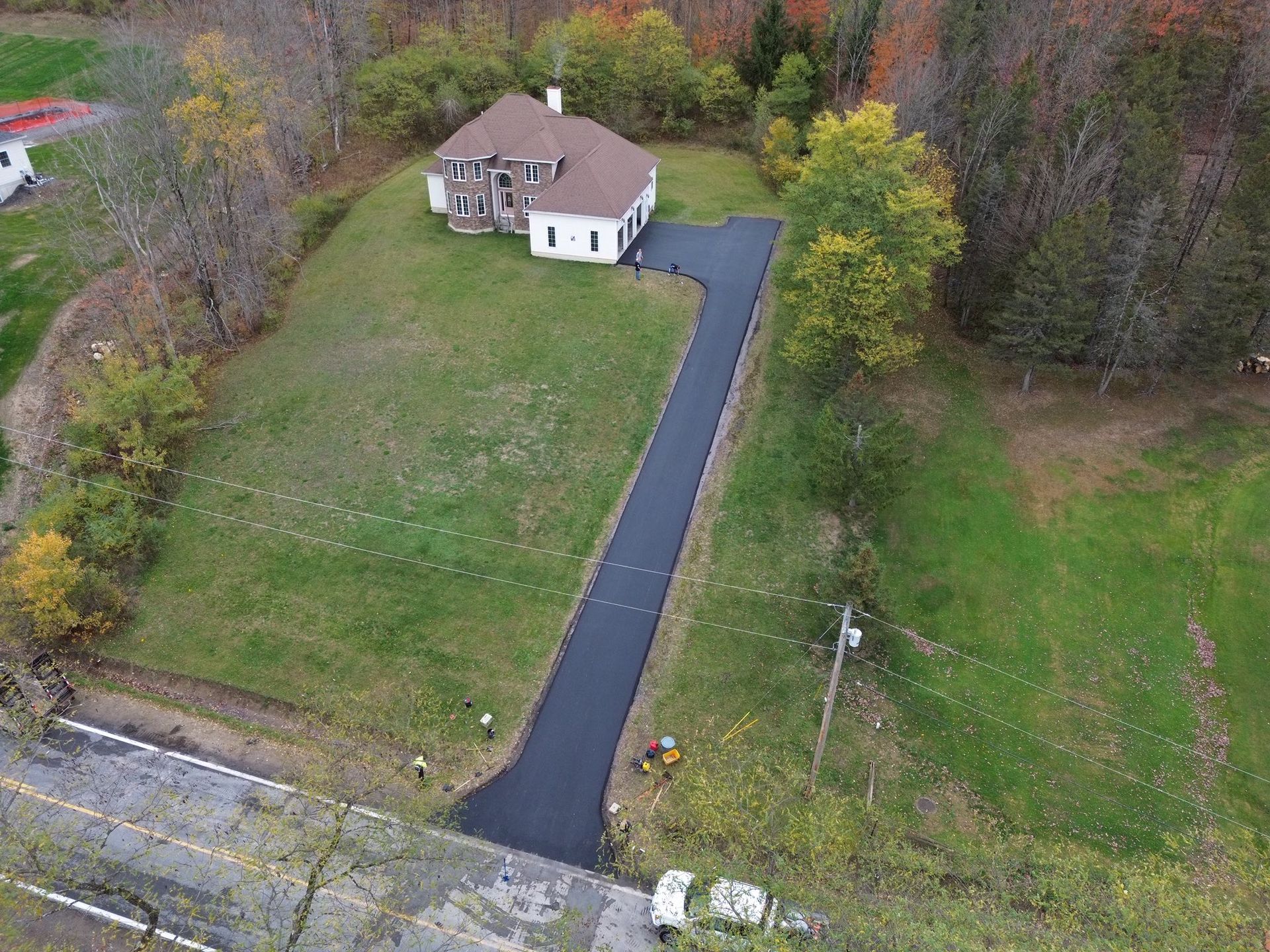 Aerial view of a house with a long black driveway, surrounded by grass and autumn trees.