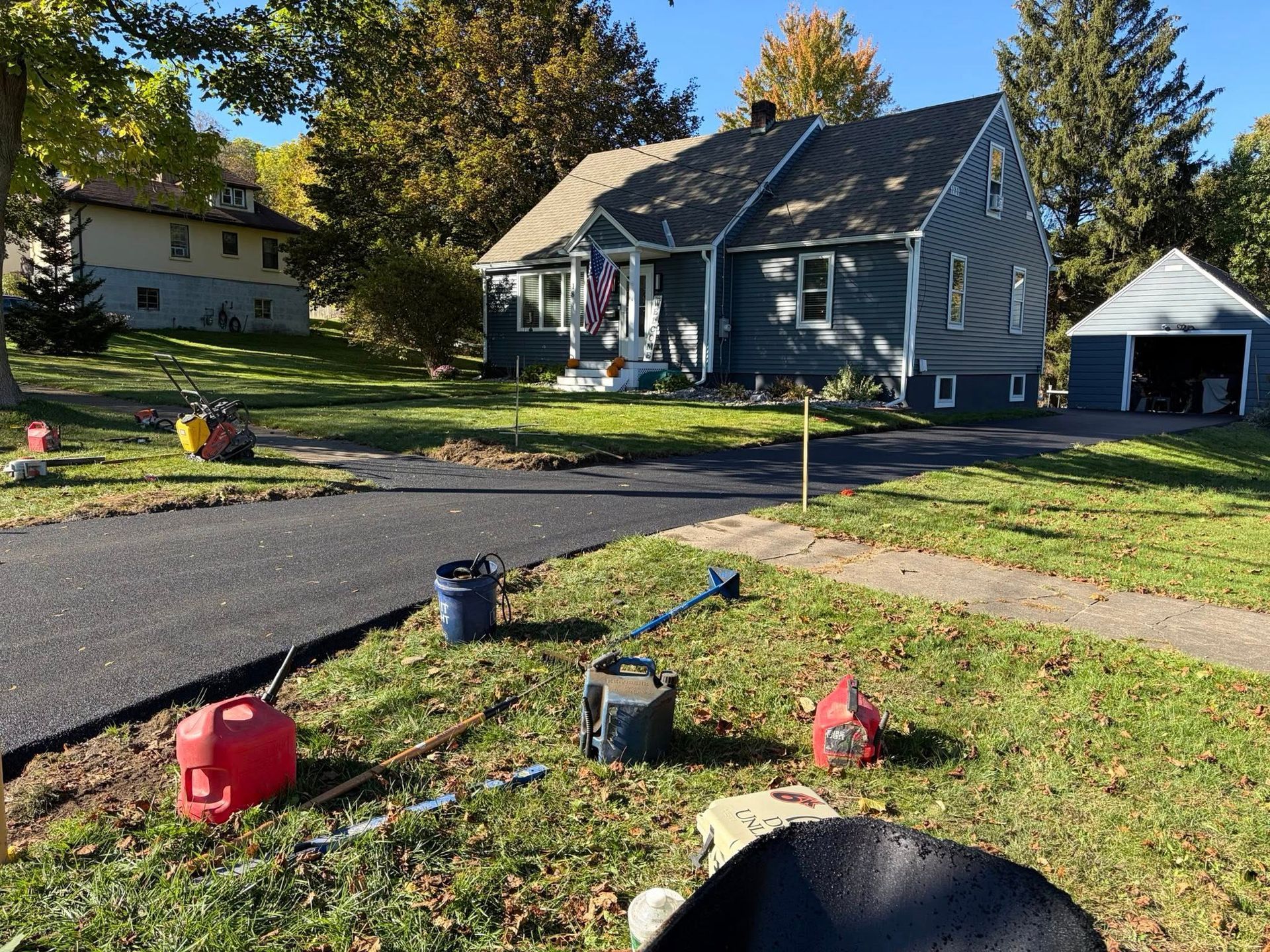 Suburban yard with a gray house, driveway, and red fuel cans on the grass under a sunny sky