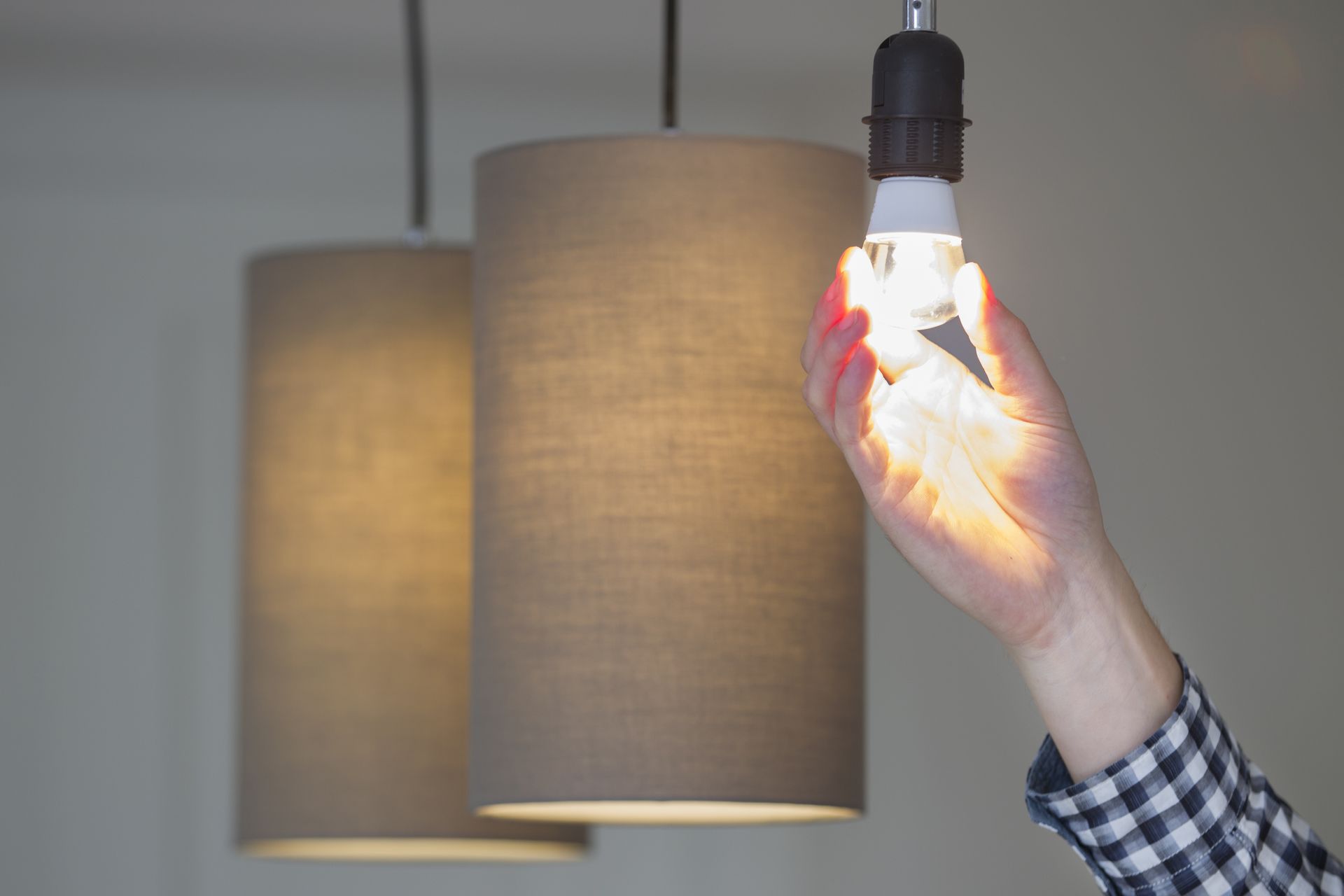 Hand holding and replacing a lit bulb in a hanging lamp, with two gray pendant shades in the background